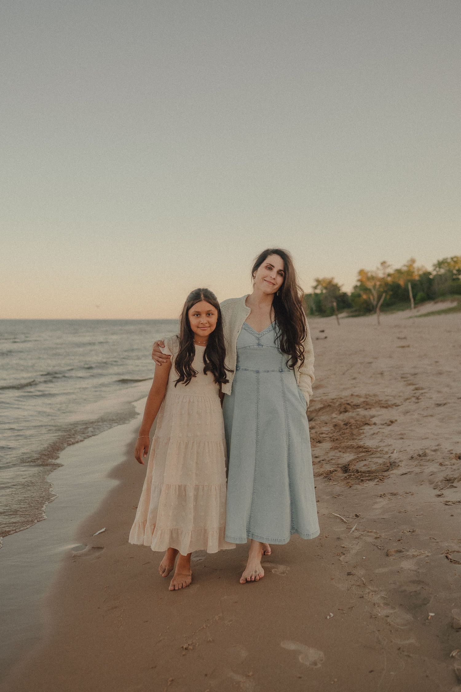 Mother and daughter in flowing dresses walk barefoot along sandy shoreline at dusk.