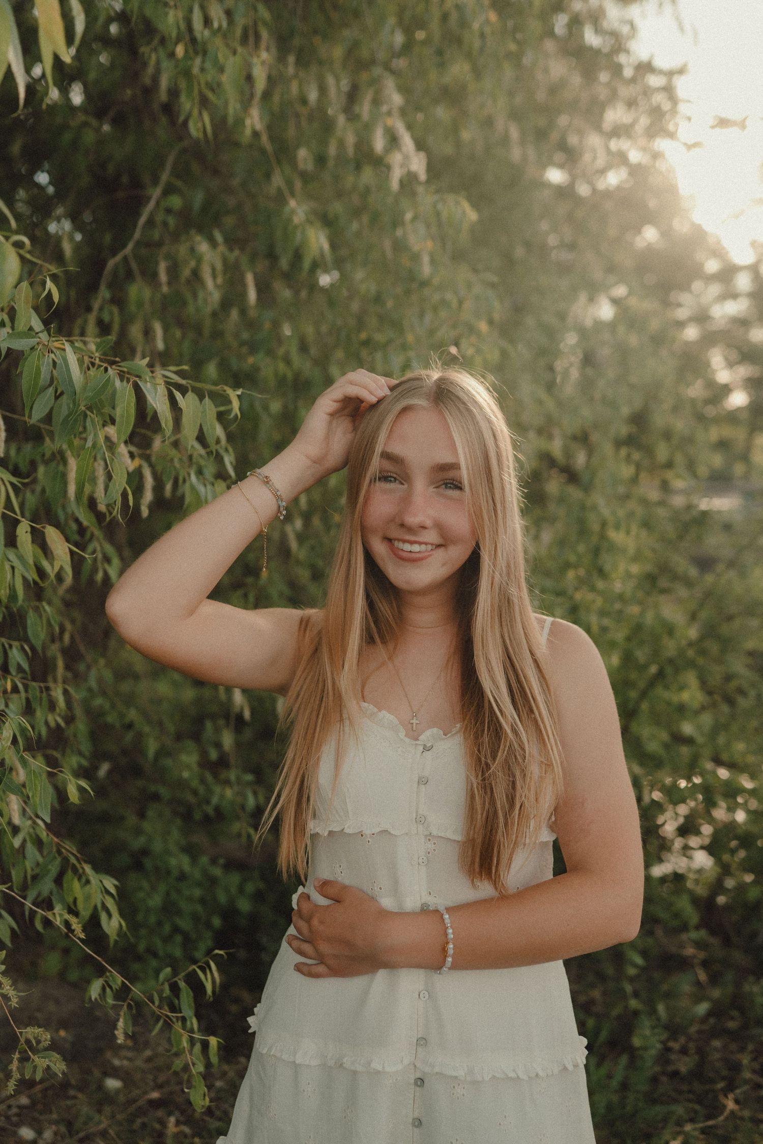 Young blonde woman in white sundress smiling outdoors among green trees in golden hour sunlight.