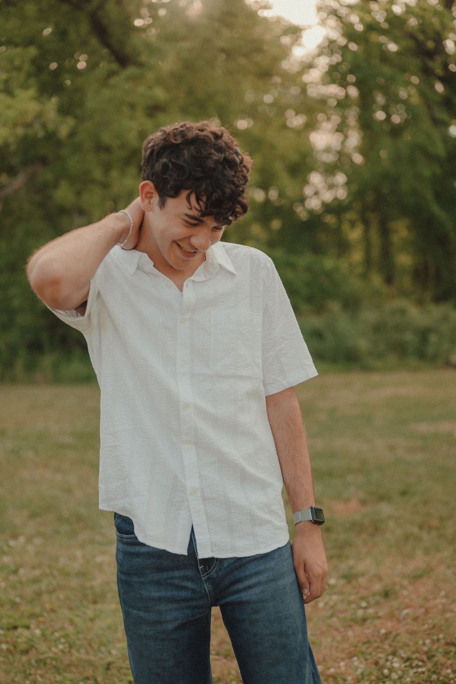 Young man with curly hair in white short-sleeve shirt and jeans smiling outdoors in a green field.