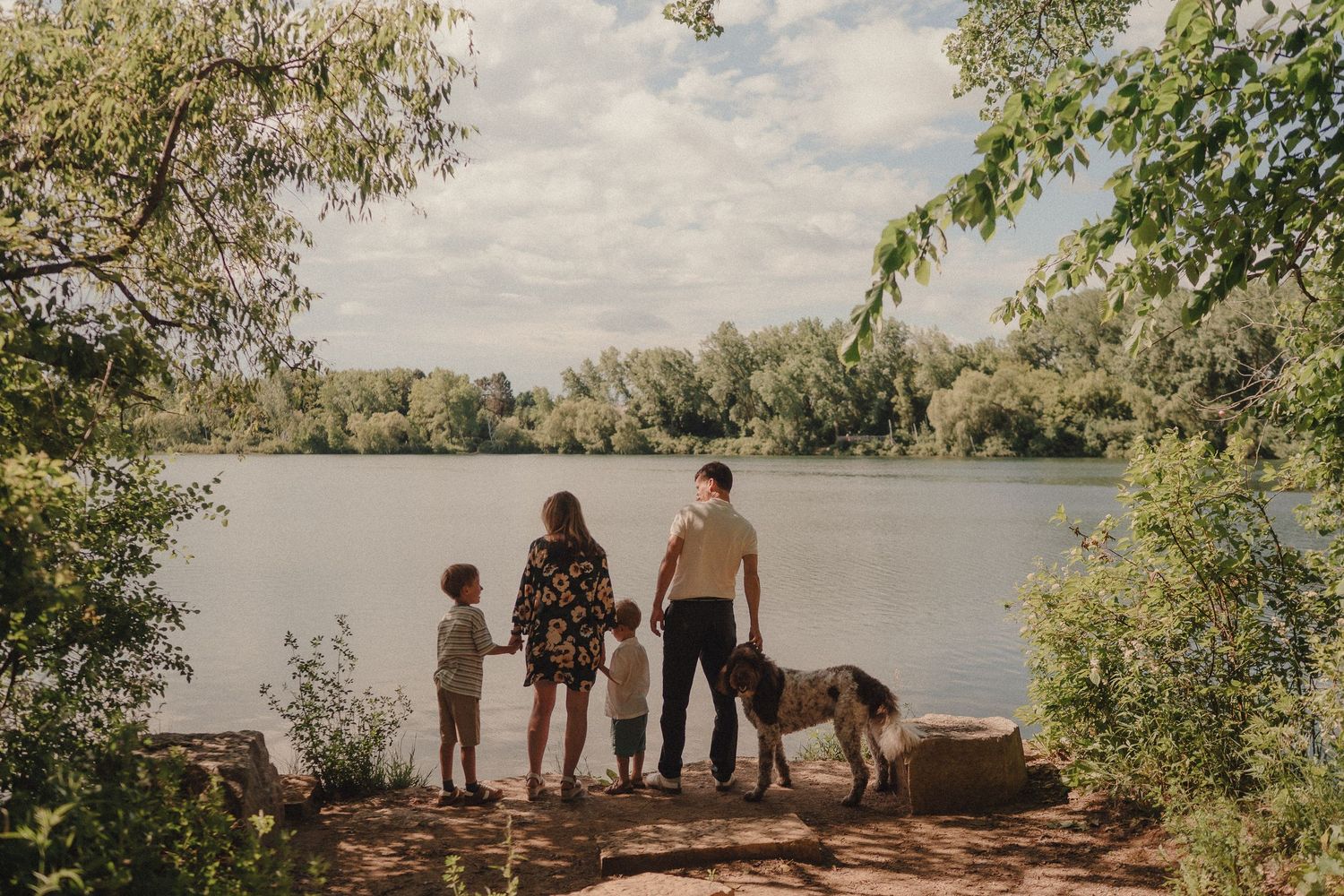 Family with child and dog standing by a serene lake surrounded by lush green trees on a sunny day.
