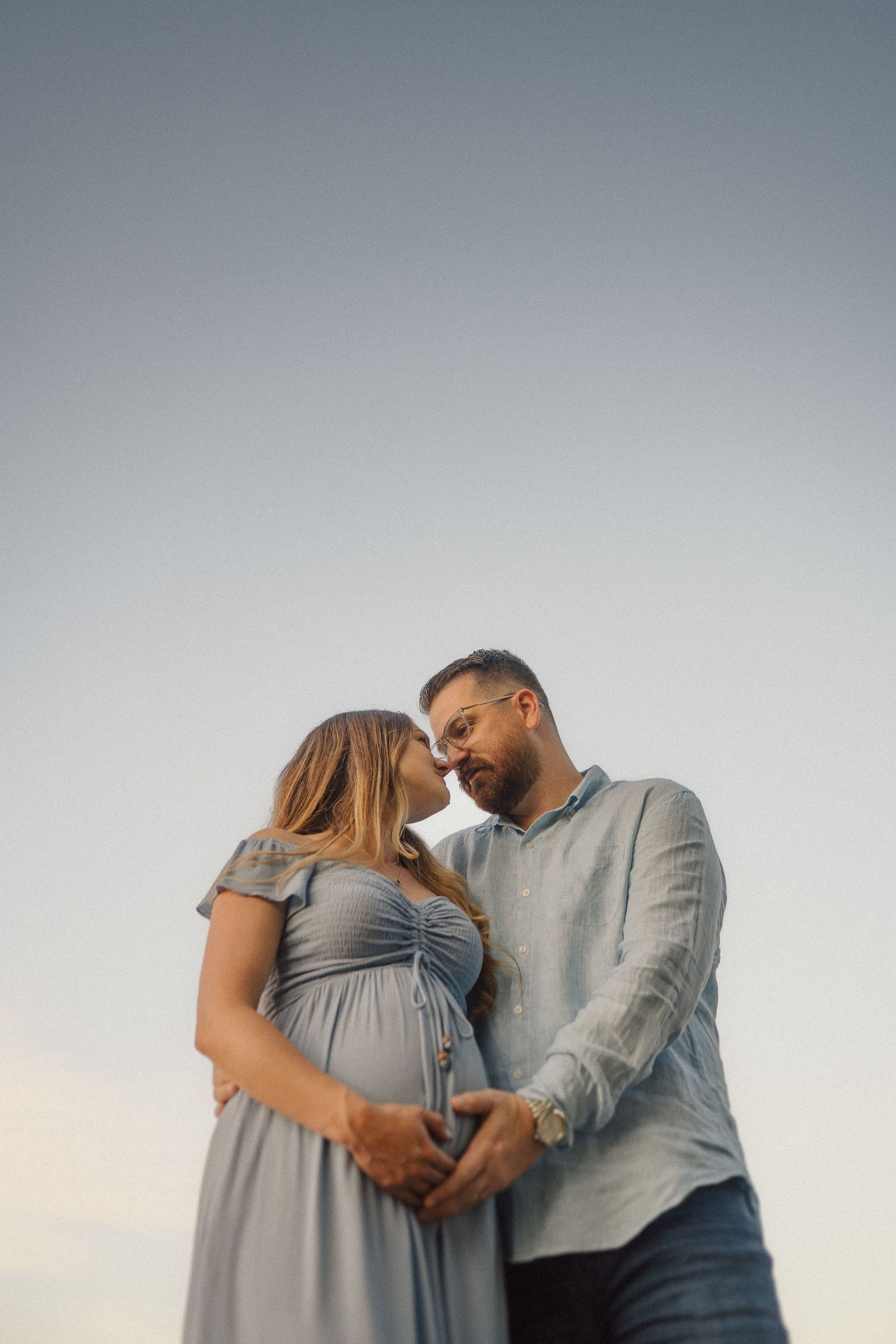A romantic maternity photoshoot featuring a couple embracing against a soft grey background at sunset.