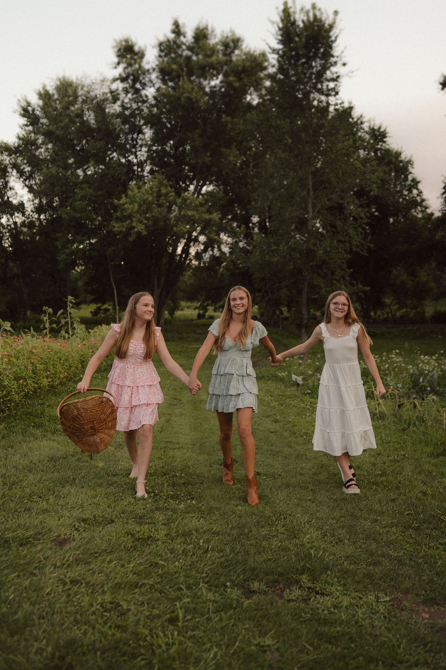 Three carefree figures in sundresses hold hands while walking through a meadow on a summer evening.