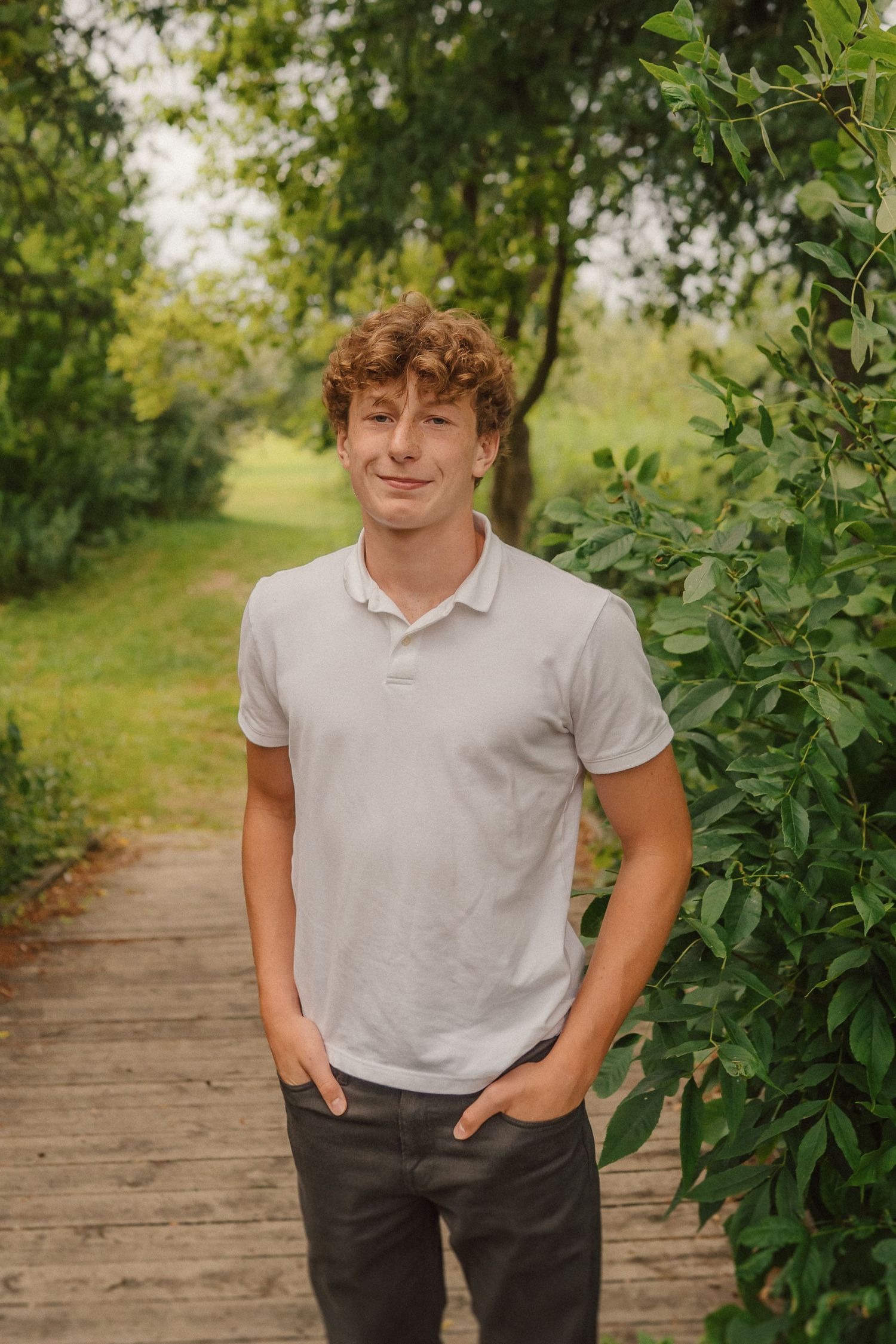A person in casual attire poses on a wooden boardwalk surrounded by leafy green foliage.