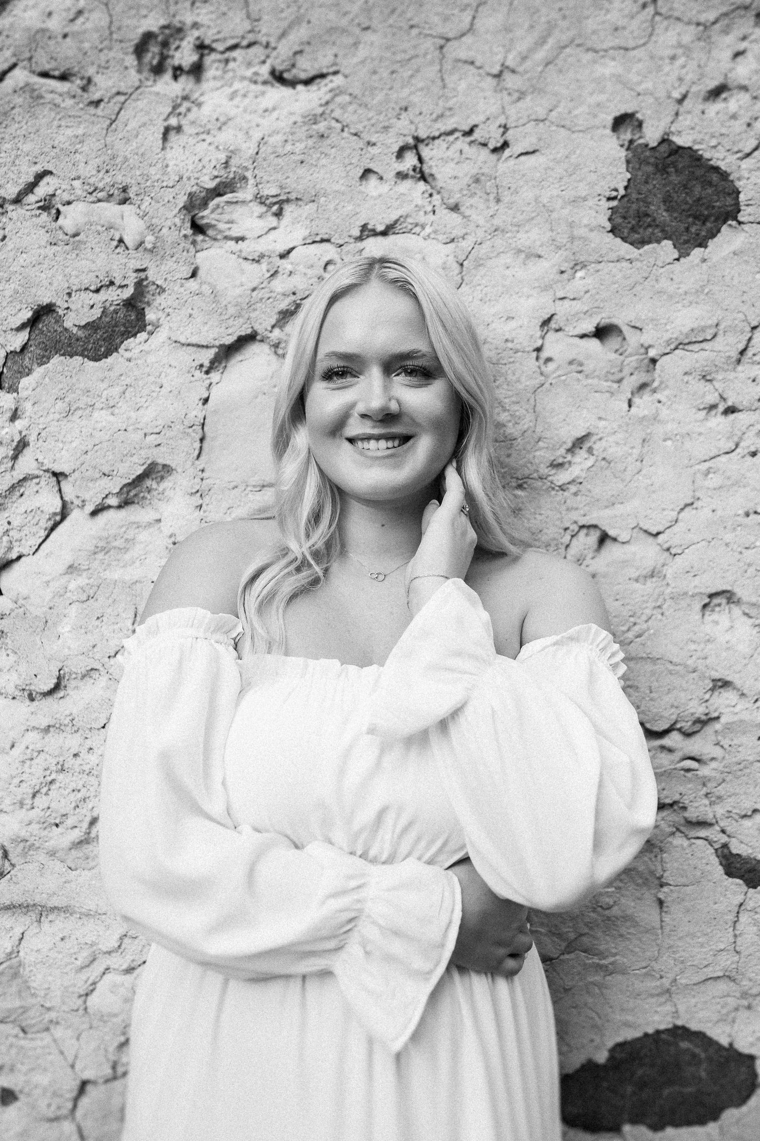 Portrait against a textured stone wall wearing a white flowing top.