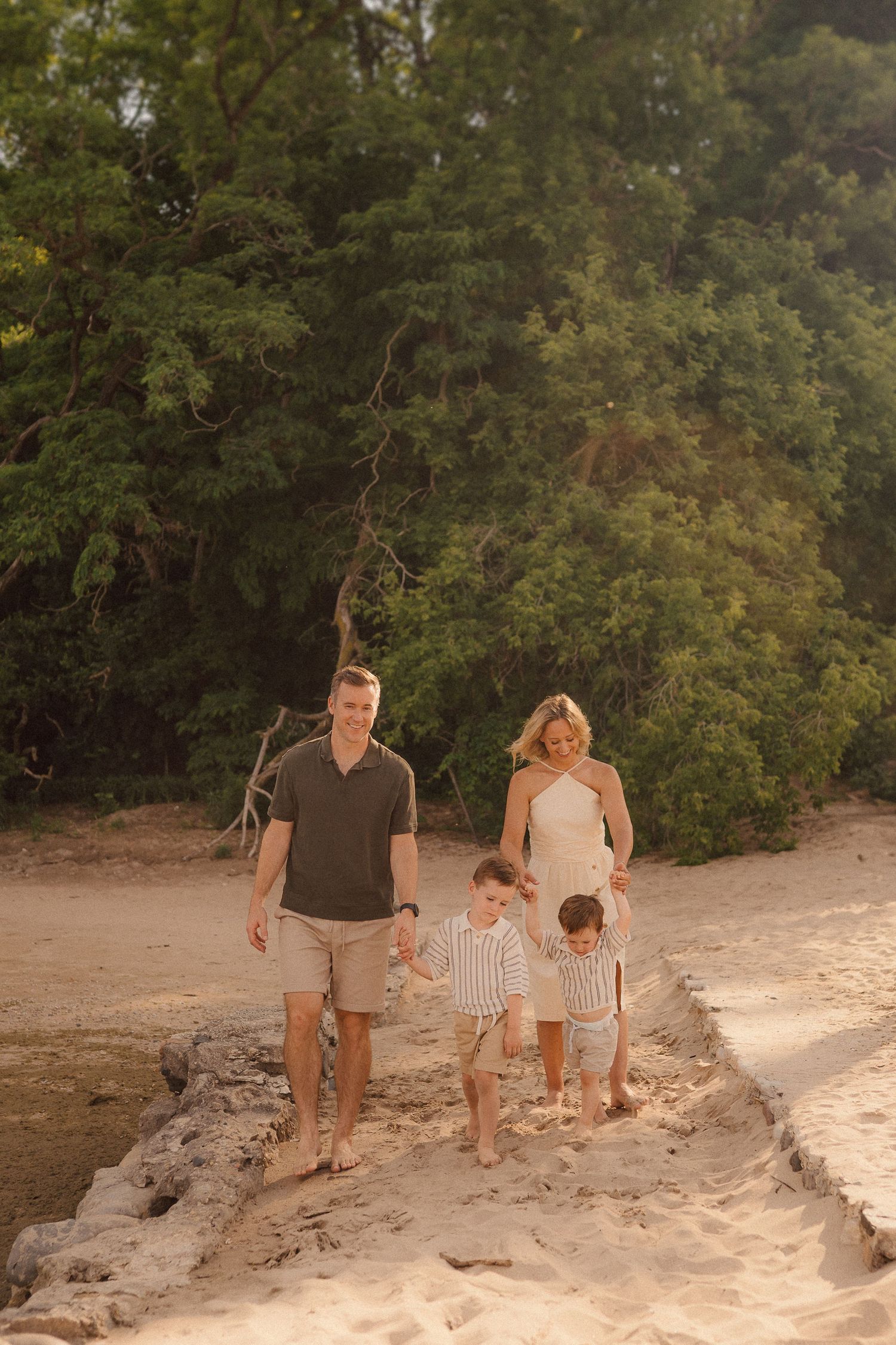 A family walks together along a sandy beach path bordered by lush green trees at sunset.