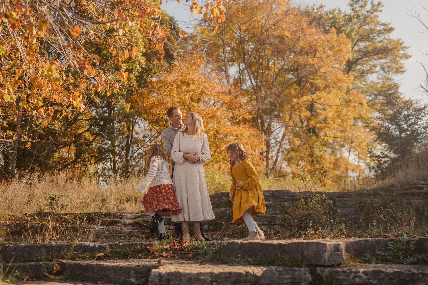 A figure stands in a white dress against vibrant autumn foliage with golden leaves and stone wall backdrop.
