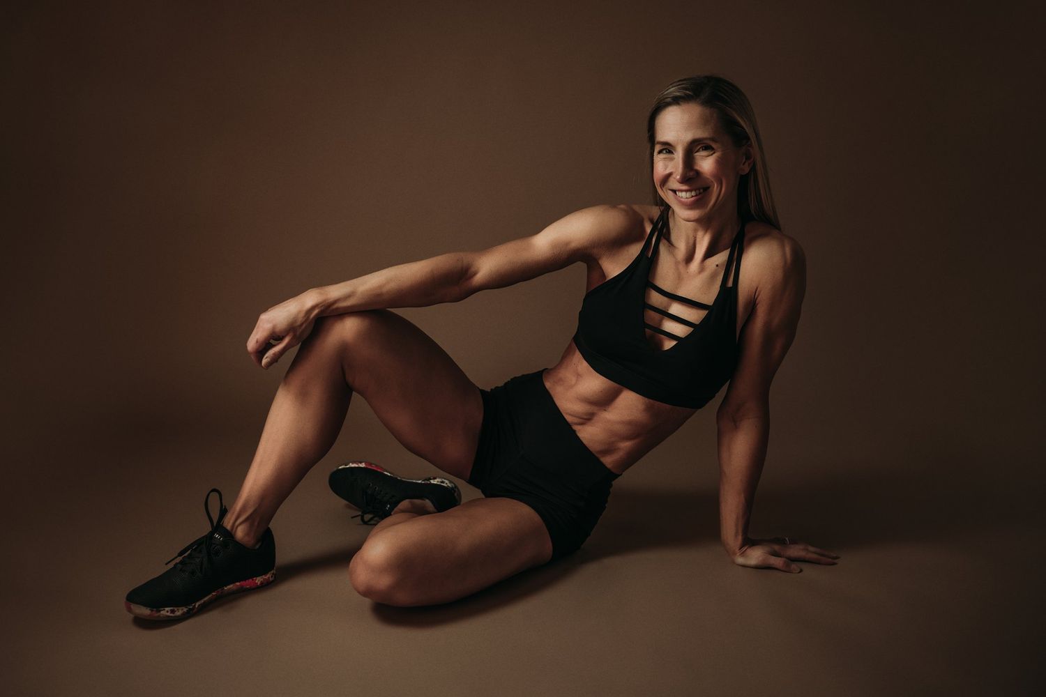 Fitness model in black activewear poses on floor against brown studio background.