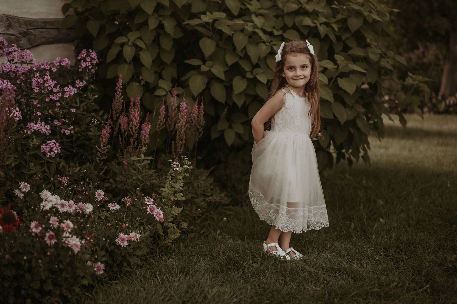 A white lace dress adorns a small figure standing near pink flowers and green vines in a garden setting.