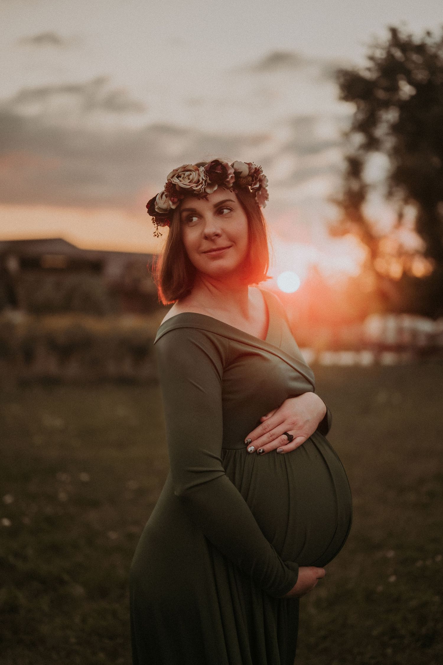 A pregnant woman wearing a floral crown poses in a green dress against a sunset sky.