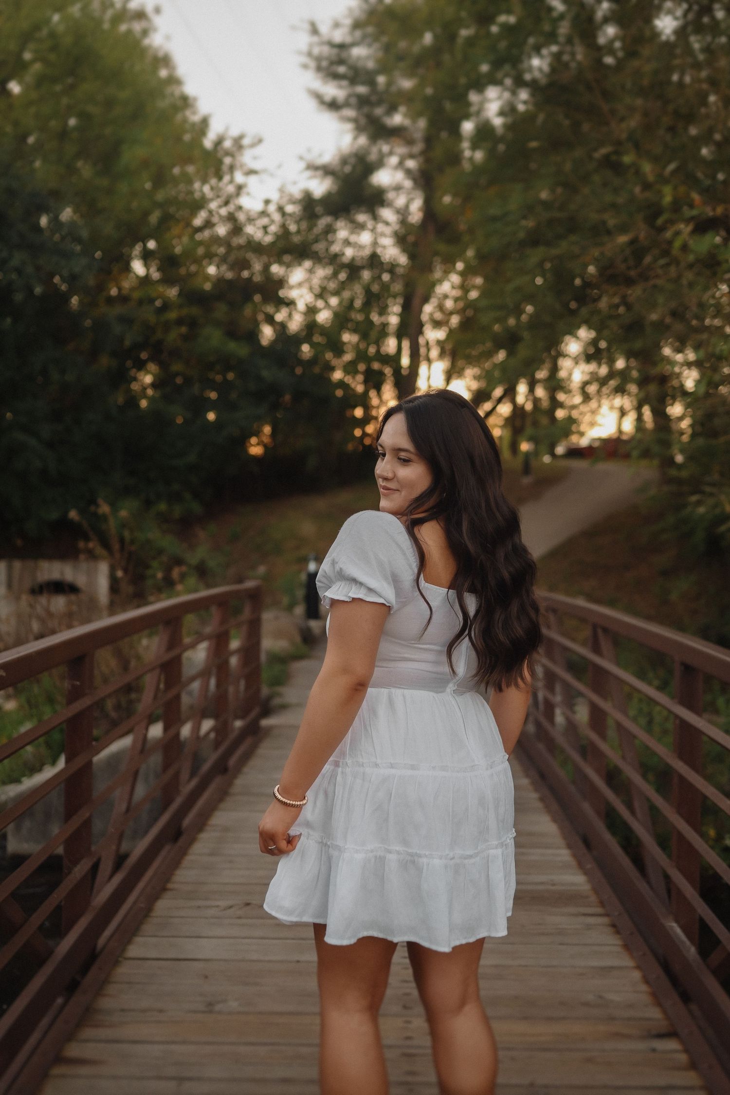 A woman in a white summer dress stands on a wooden bridge during golden hour in a forest setting.