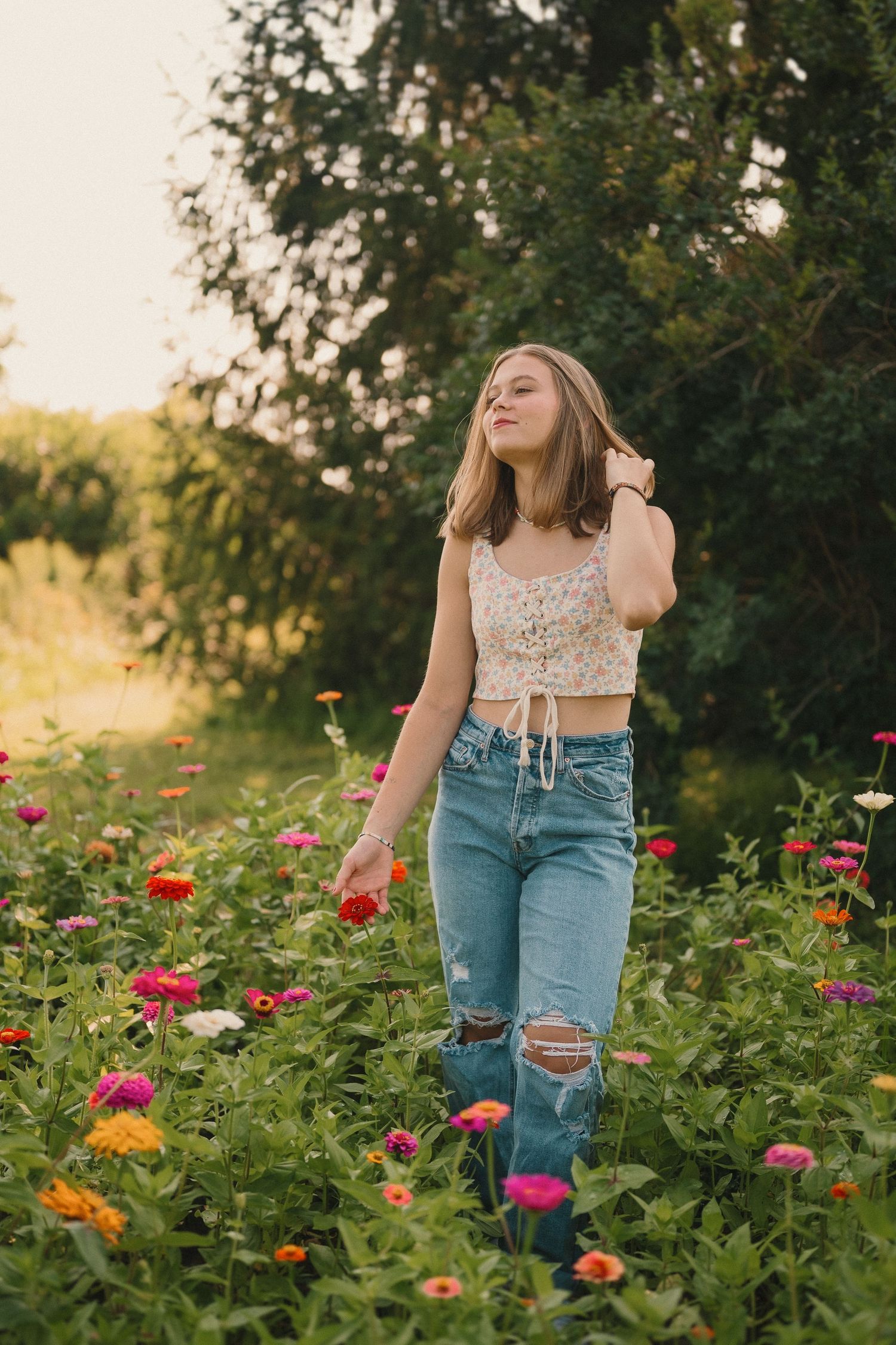 Young woman in crop top and ripped jeans stands among colorful wildflowers in a garden at sunset.