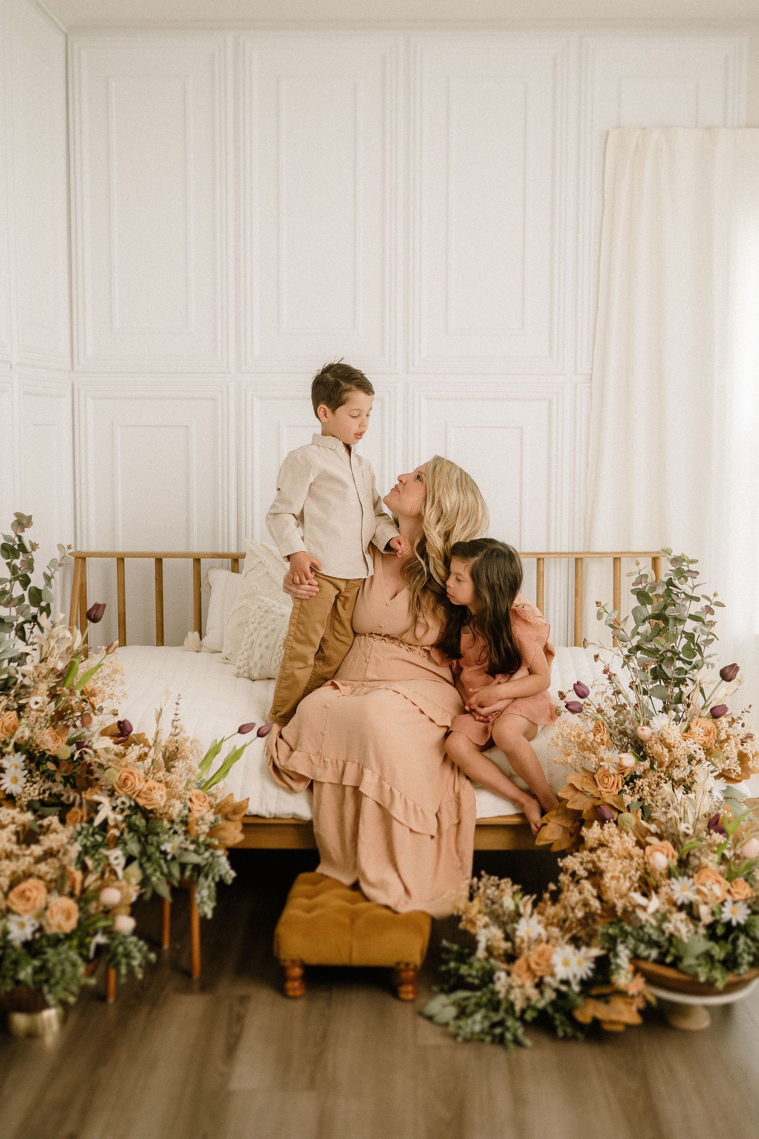 Family portrait in white room with rustic wood bed frame surrounded by peach and pink floral arrangements.