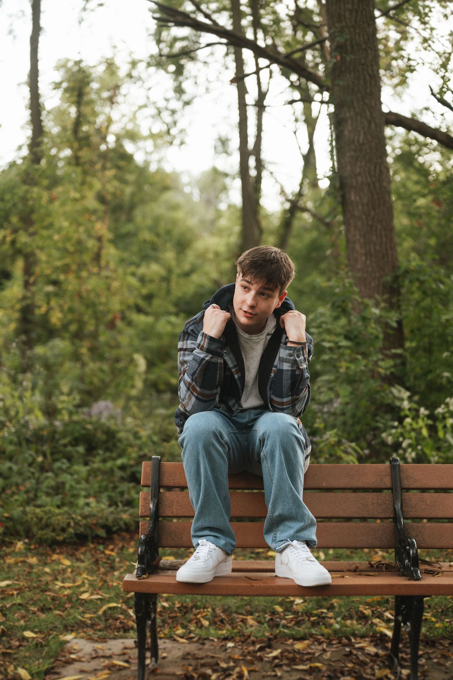 Person sitting alone on wooden park bench in autumn forest wearing jeans and white sneakers.
