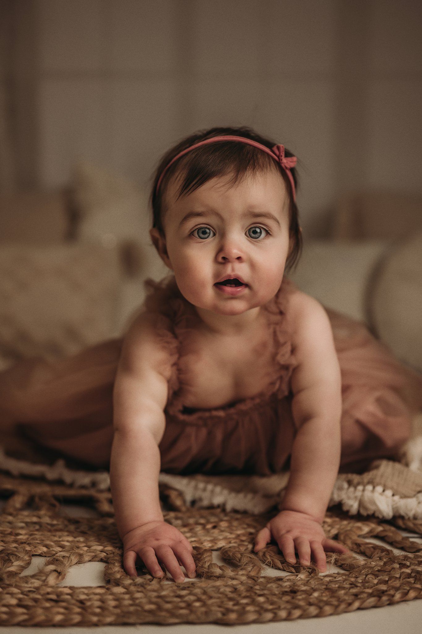 Baby wearing pink tulle dress crawling on jute rug in soft-lit indoor setting.