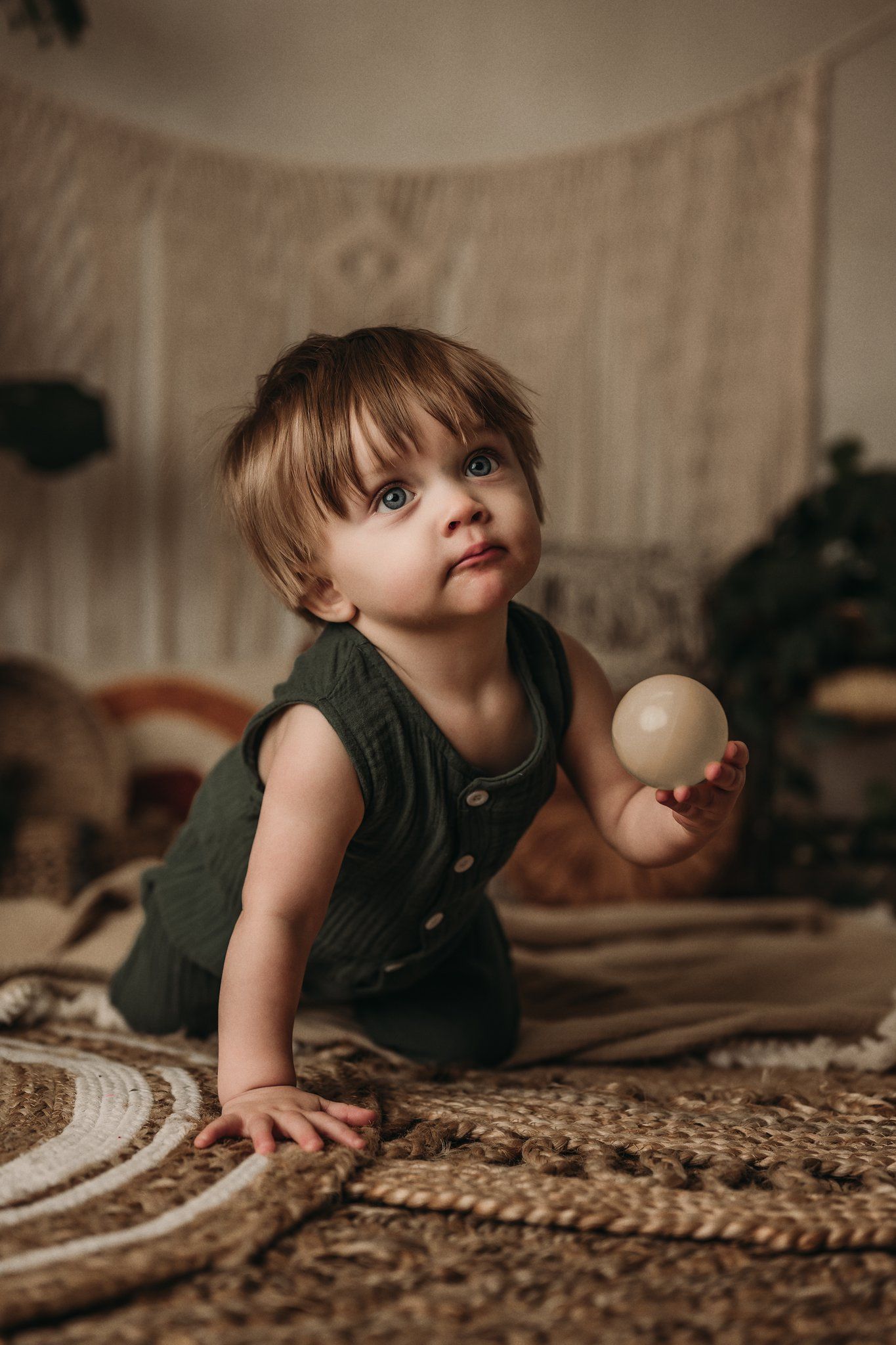 Child in green romper plays with wooden ball on textured rug with macrame backdrop.