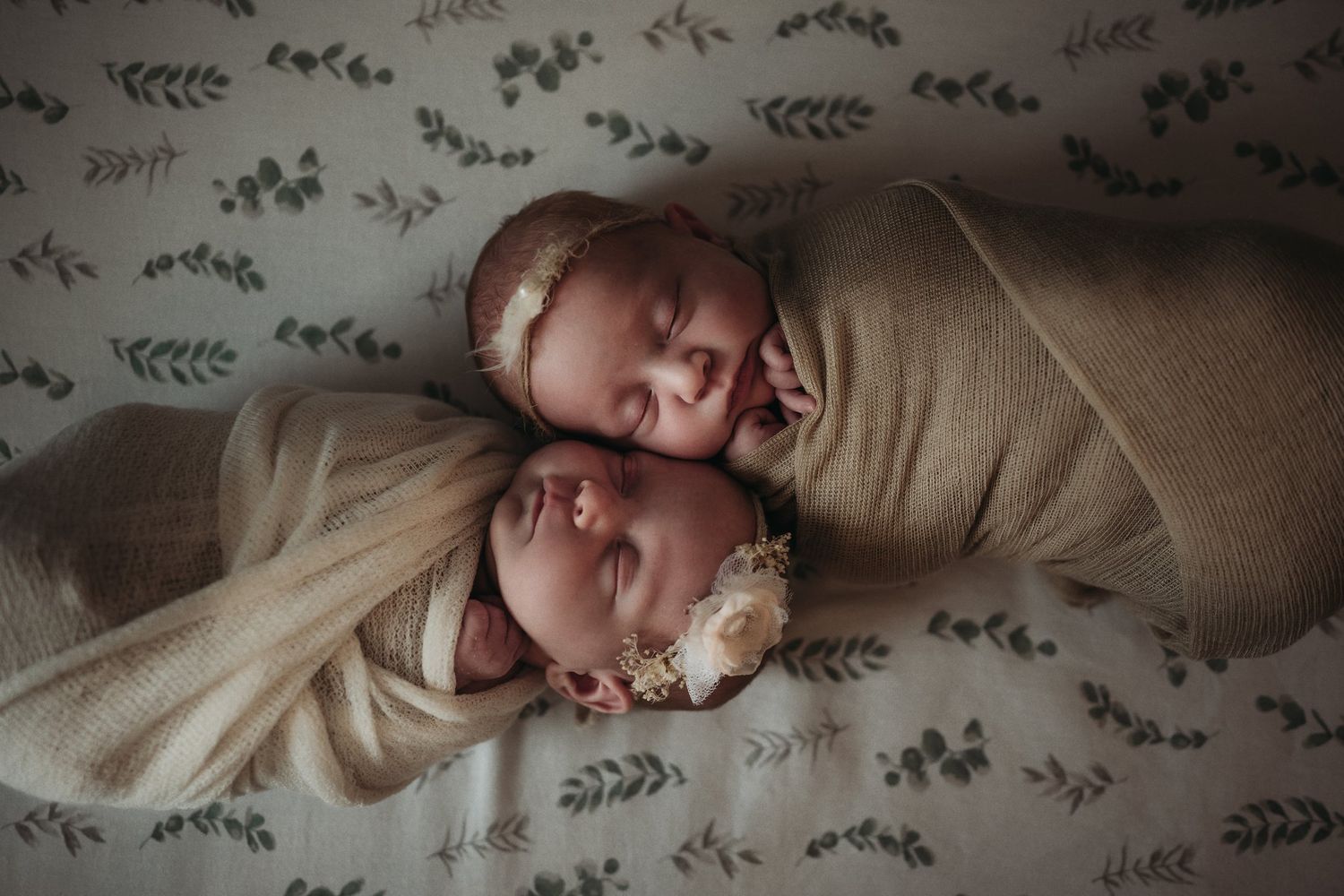 Twin babies wrapped in beige swaddles sleeping peacefully on floral pattern bedding.
