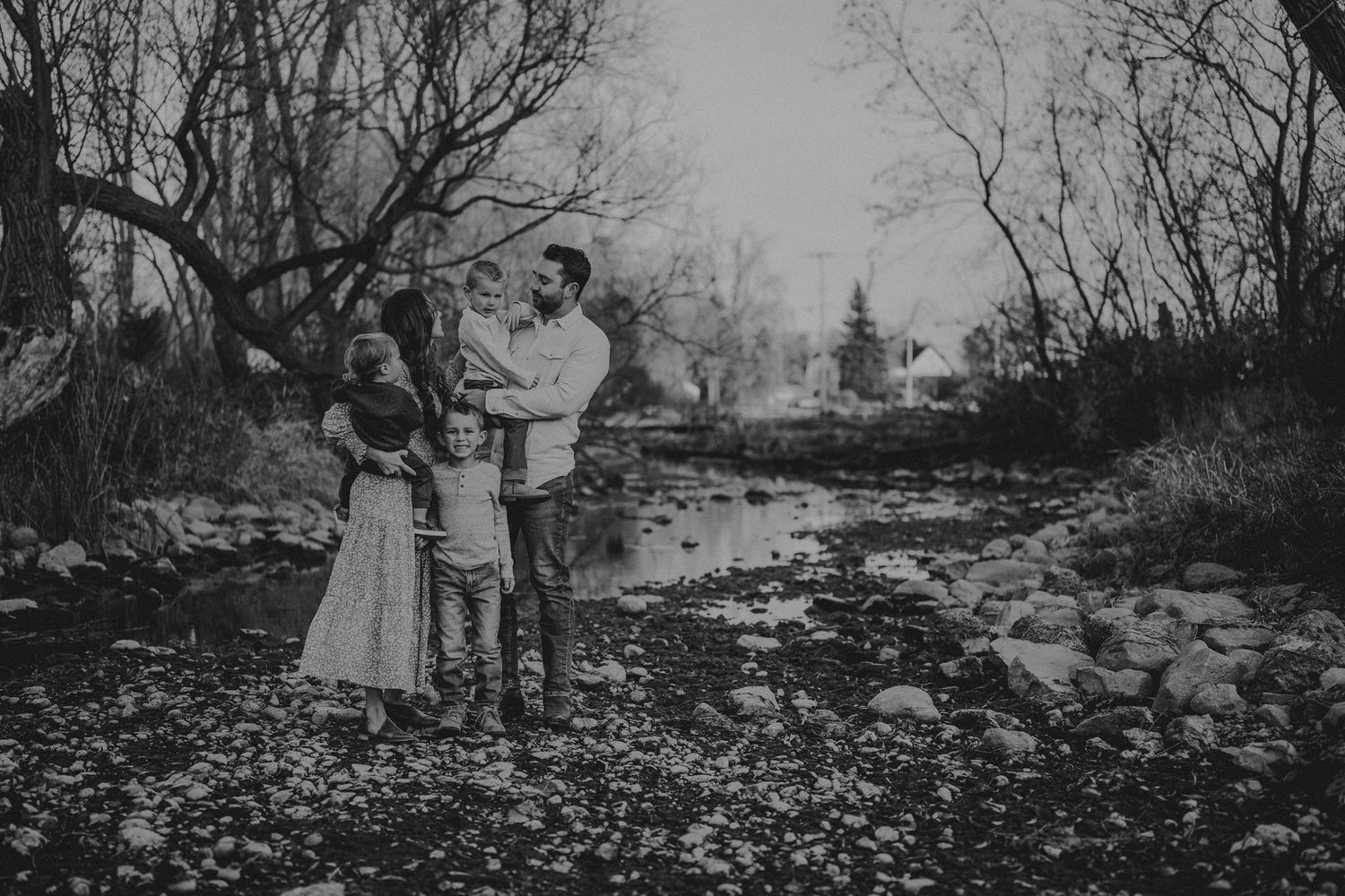 Black and white family portrait beside stream with bare trees and church spire visible in distance.