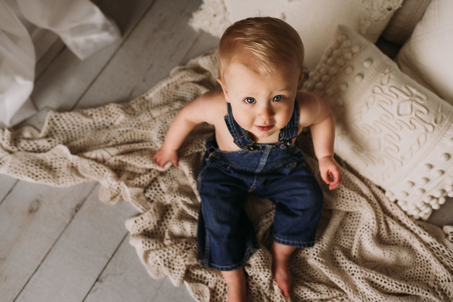 A small child in blue overalls rests on a knit blanket on a wooden floor.