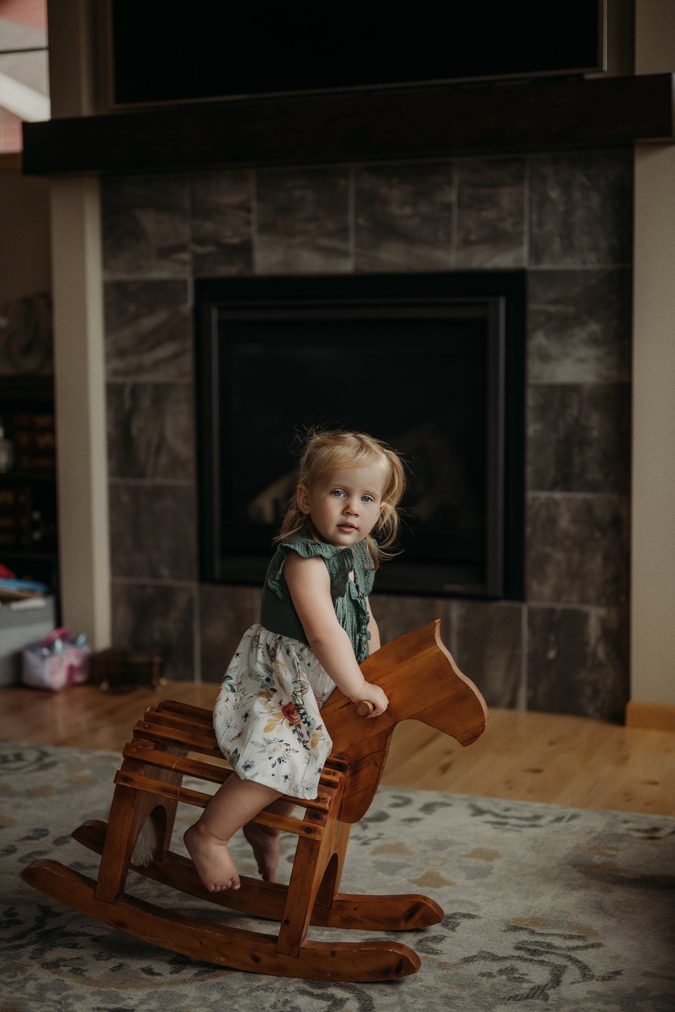 A toddler in a patterned dress rides a wooden rocking horse in front of a stone fireplace.