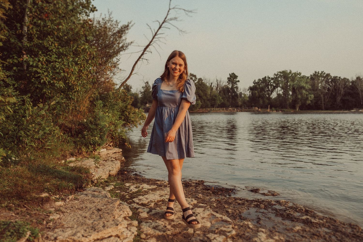 Someone wearing a blue dress stands on a rocky shoreline during golden hour.