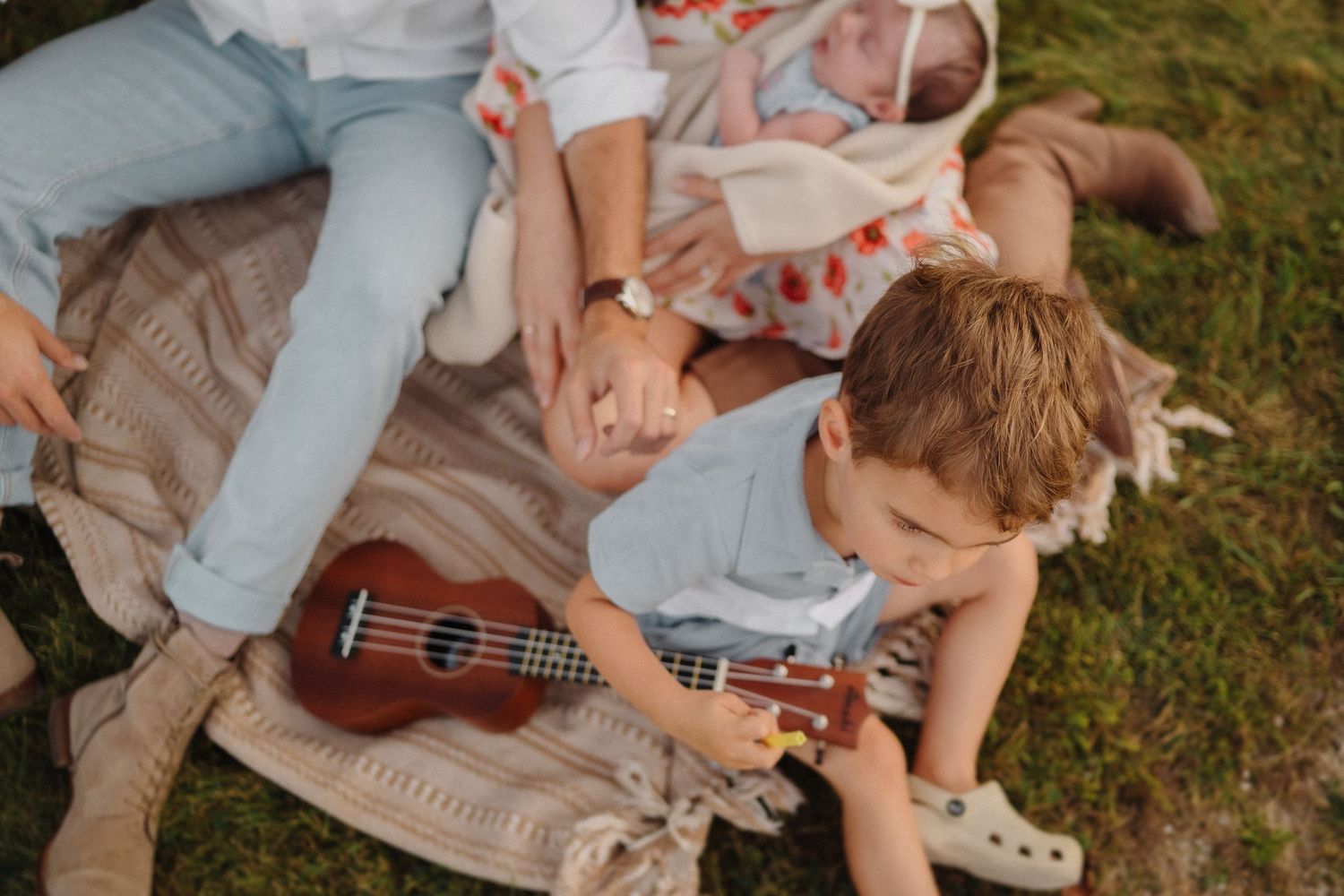 A child playing with a ukulele while relaxing on a picnic blanket outdoors.