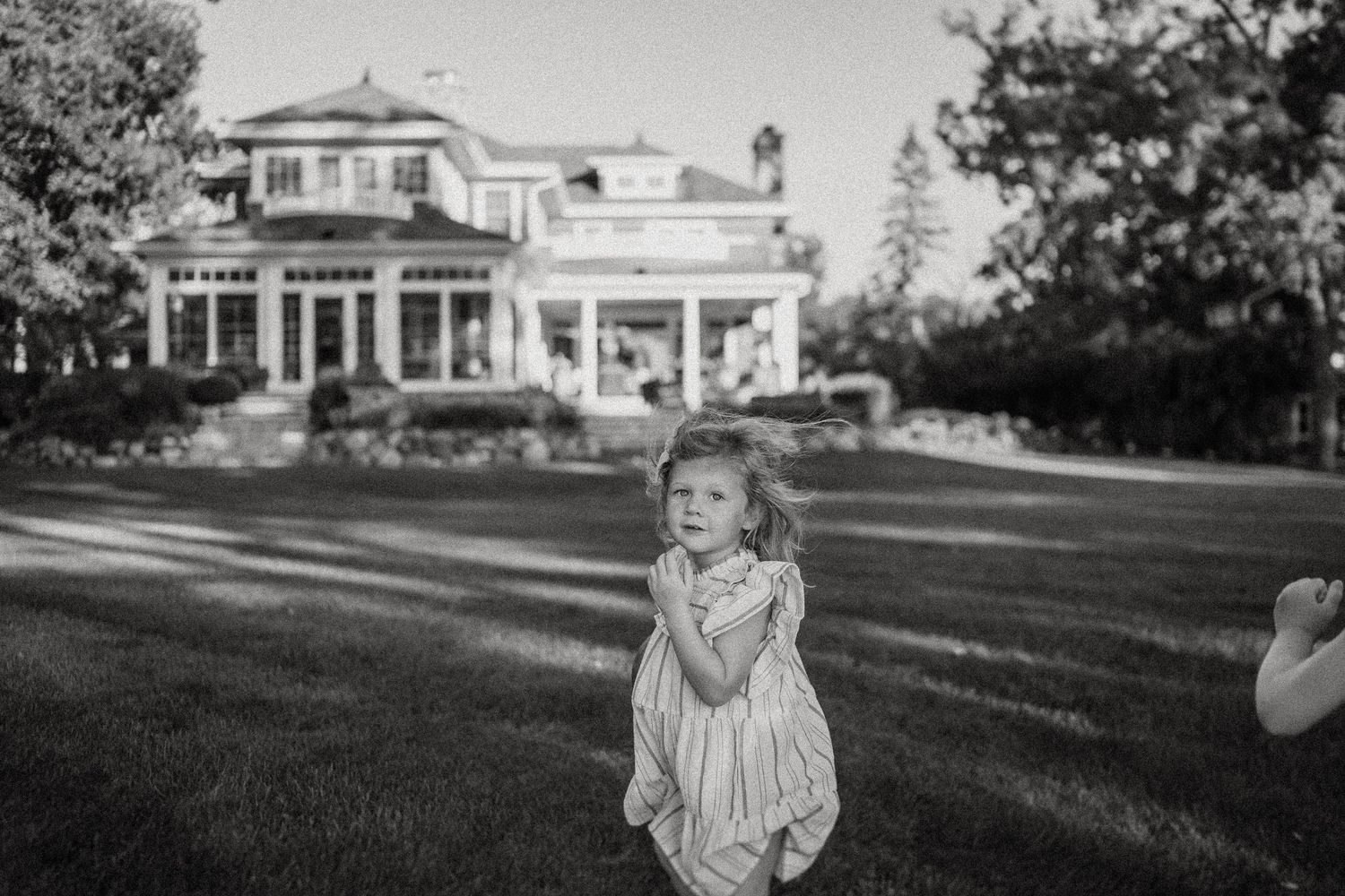 A young child in a white dress plays on a lawn in front of a large estate house.