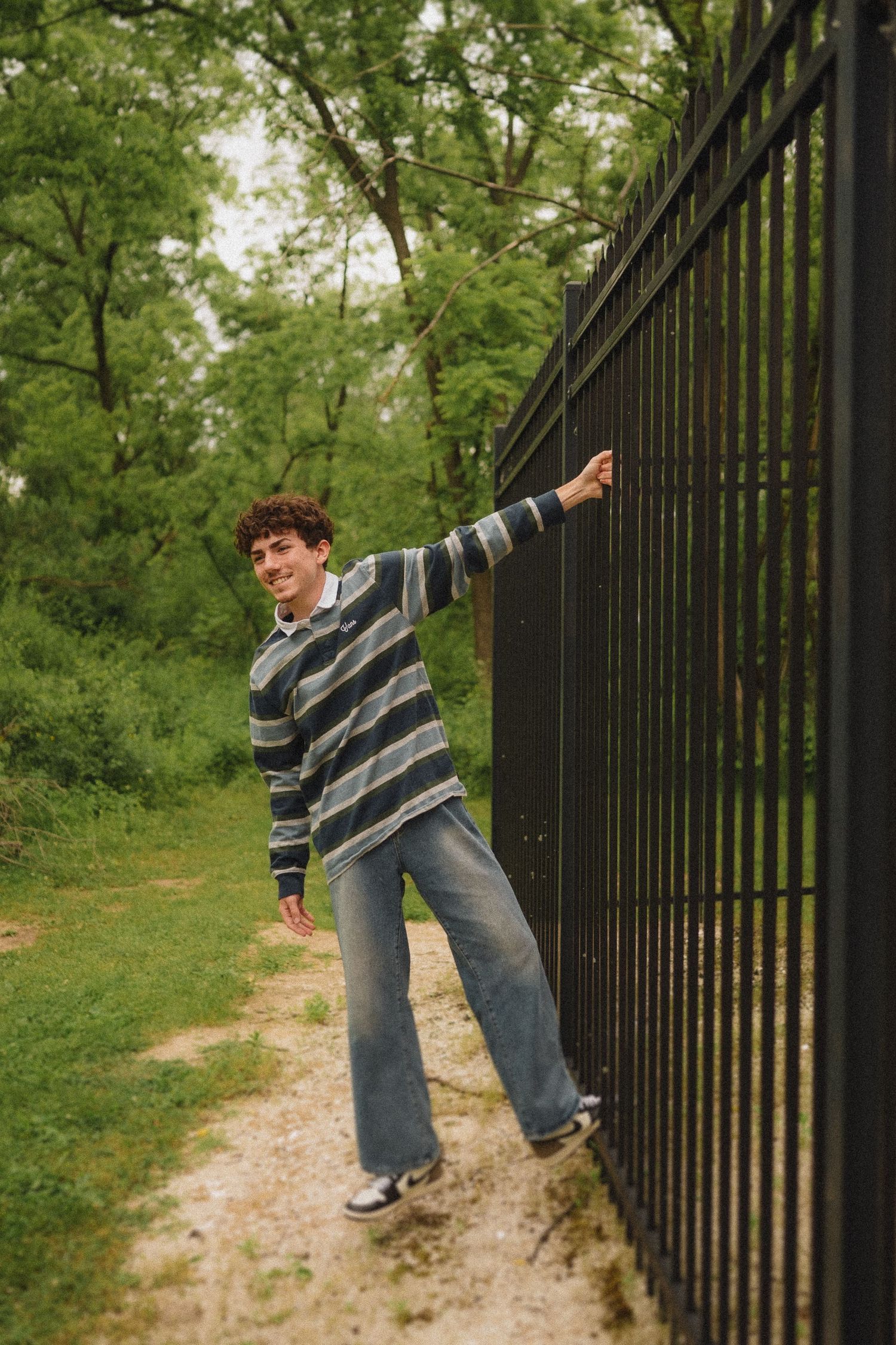Person in striped sweater leans playfully against a black iron fence along a dirt path surrounded by green trees.
