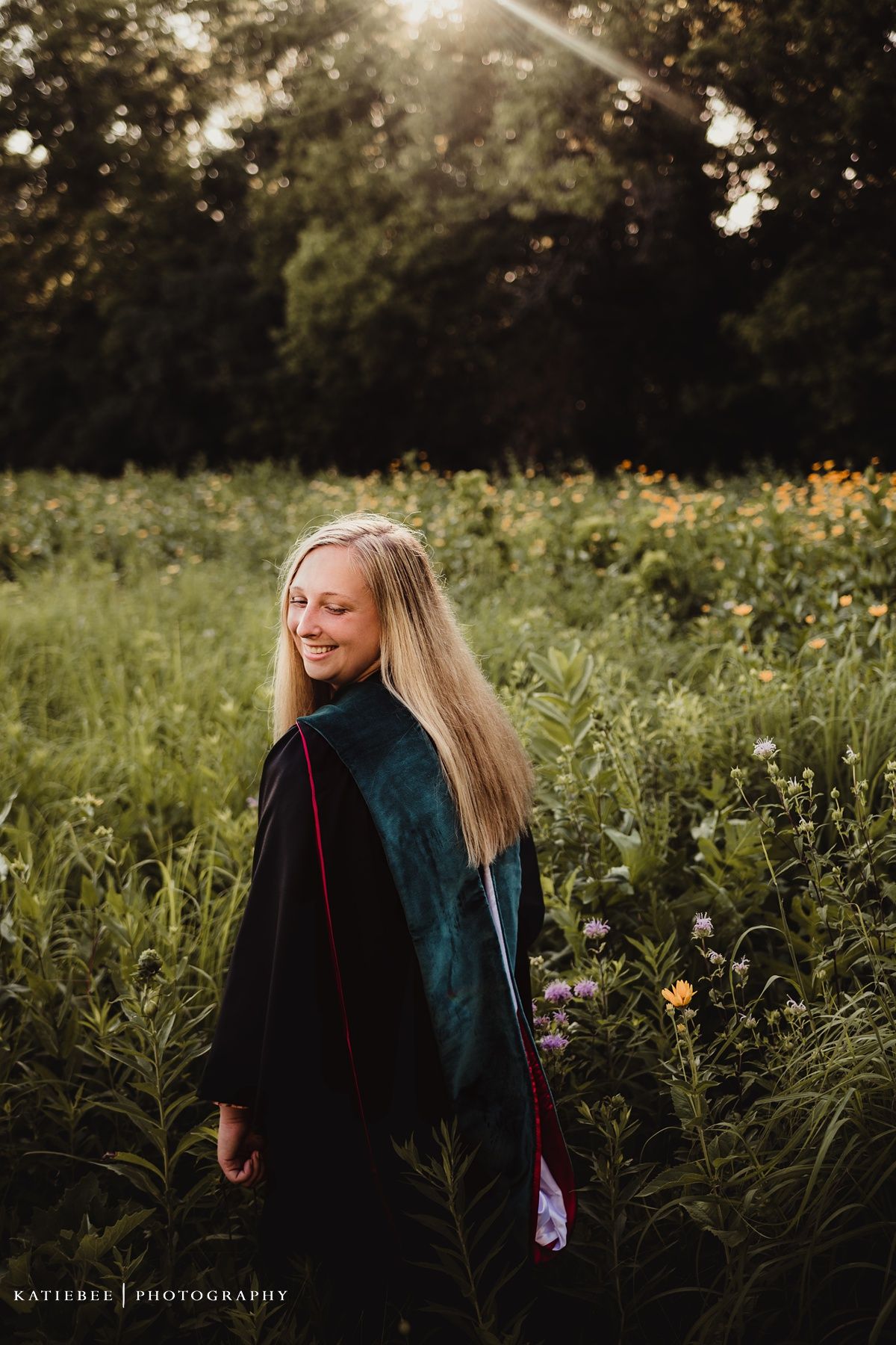 Woman in graduation gown turns in a golden meadow during sunset.