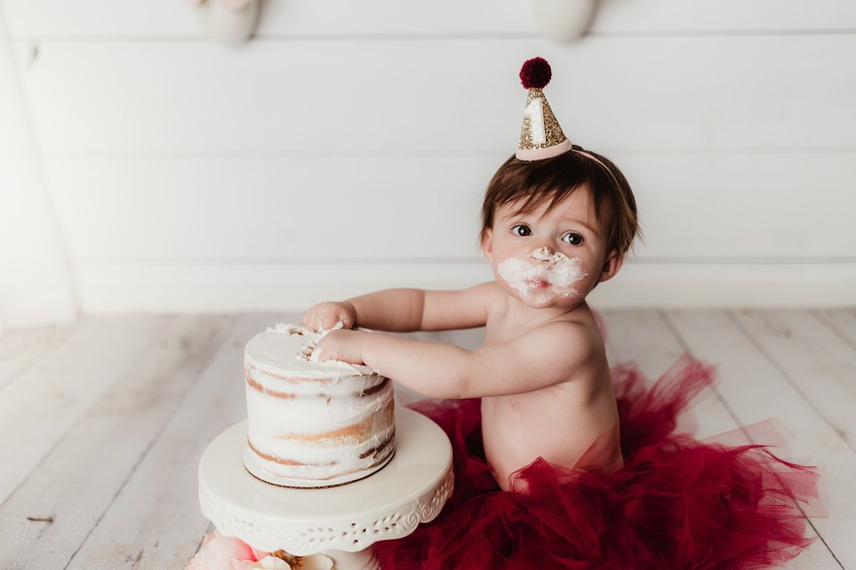 Baby in burgundy tutu touching white layered birthday cake during photo shoot.