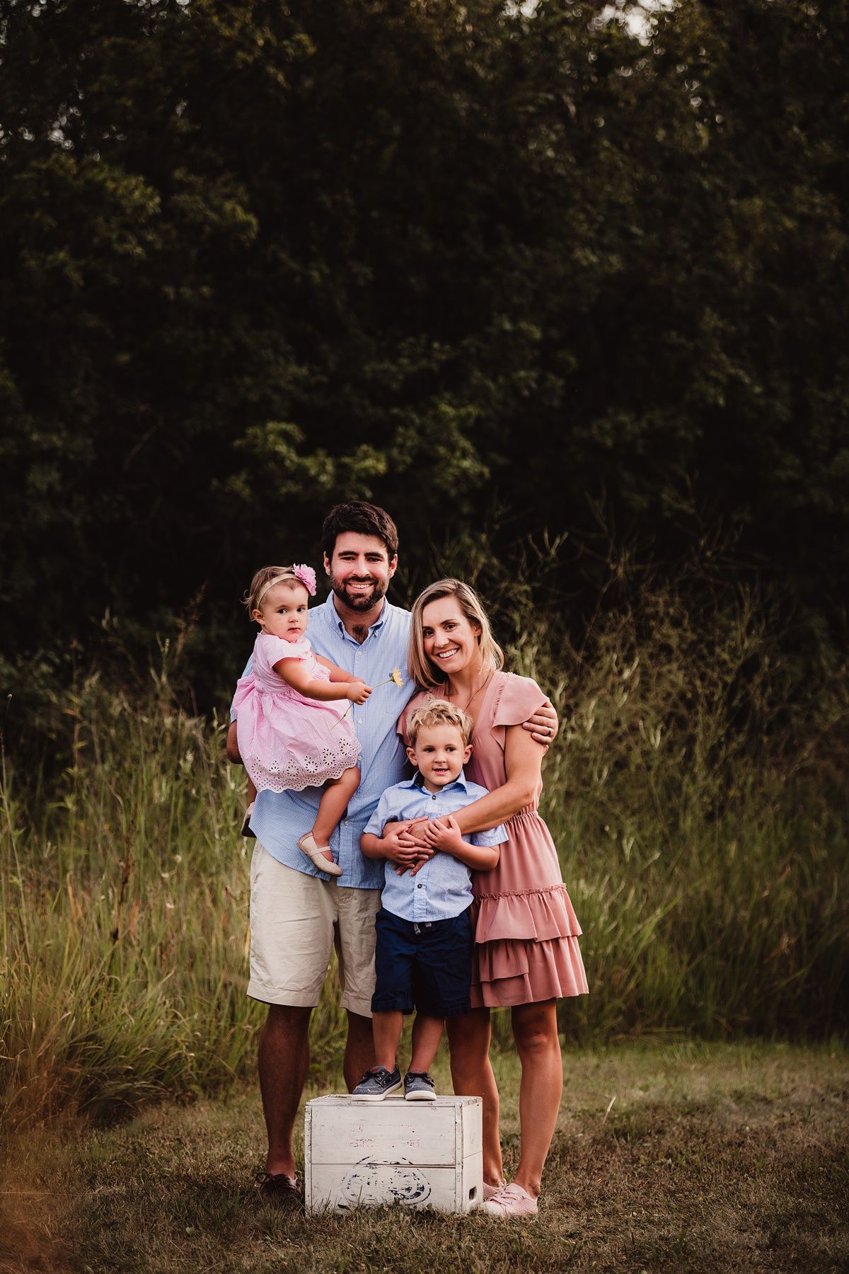 A family of four poses together on a wooden crate in a rustic outdoor setting.