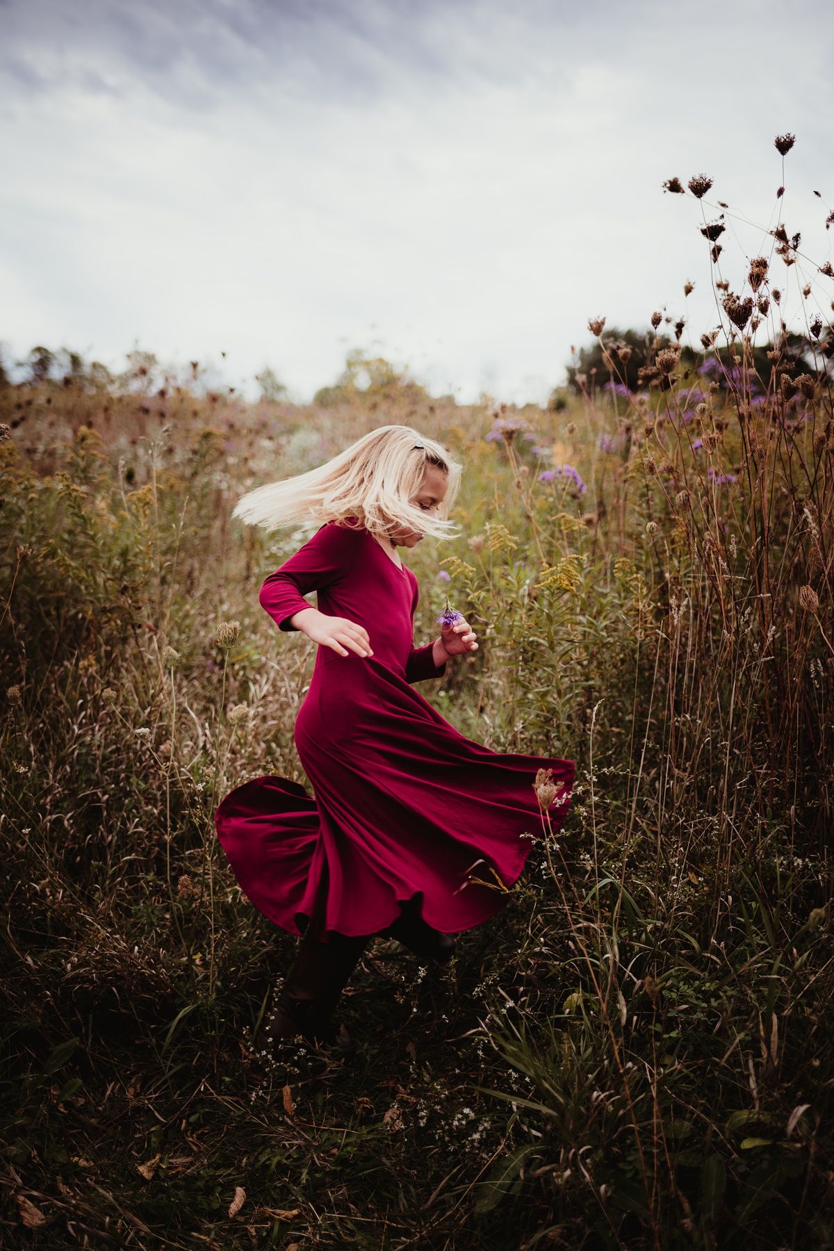 Woman in flowing red dress twirling through a wildflower meadow under cloudy skies.