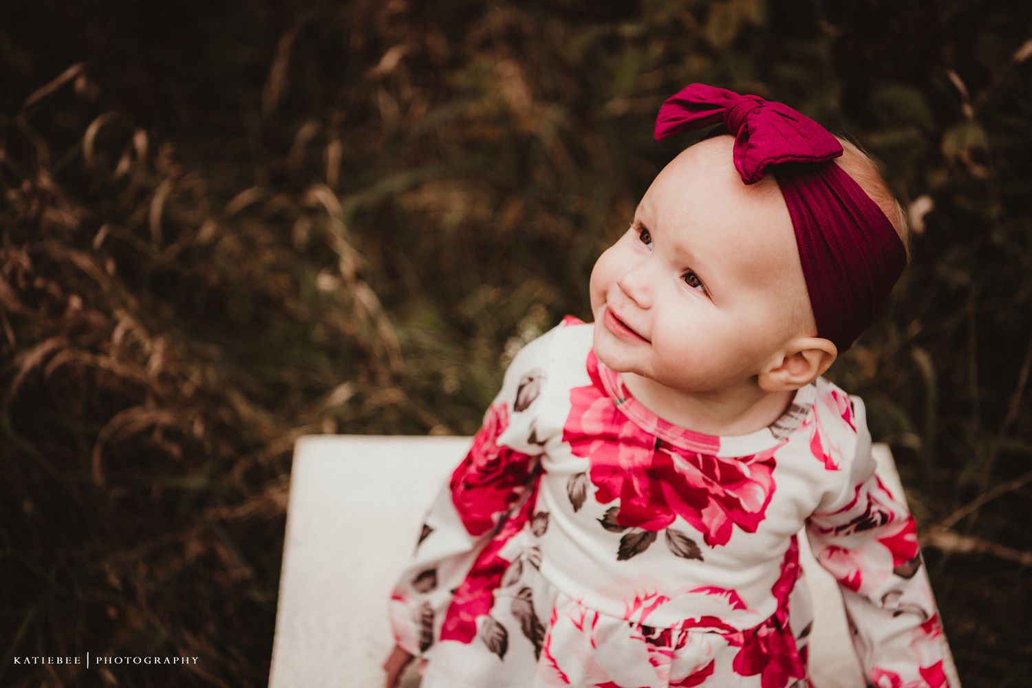 Baby wearing a floral dress and red bow headband smiles sweetly.