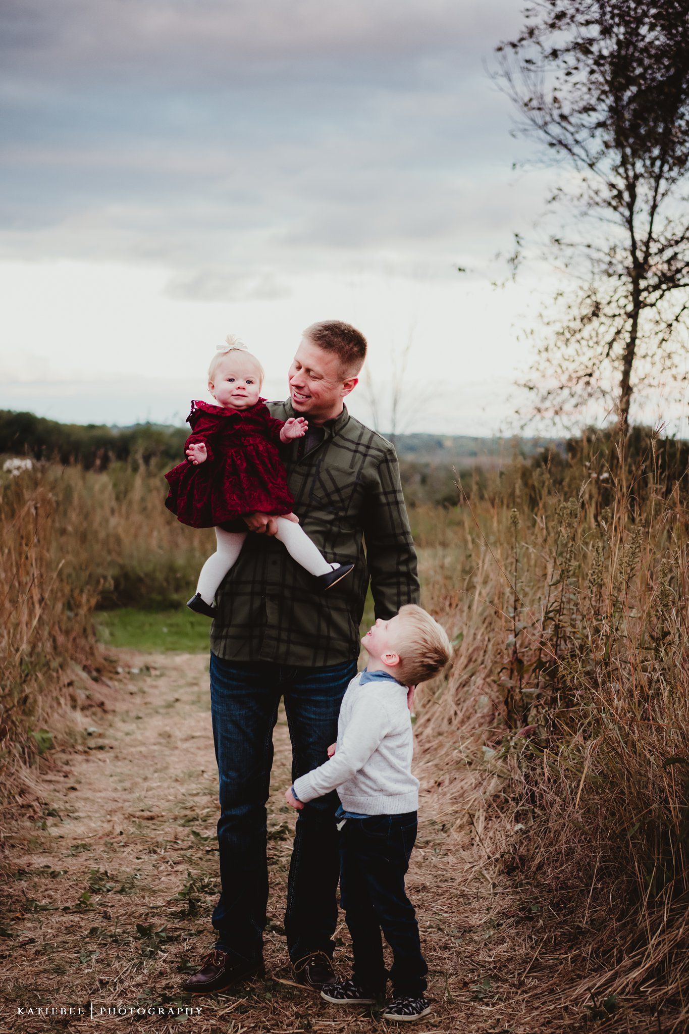 A heartwarming outdoor family portrait taken on a dirt path surrounded by tall grass and moody skies.