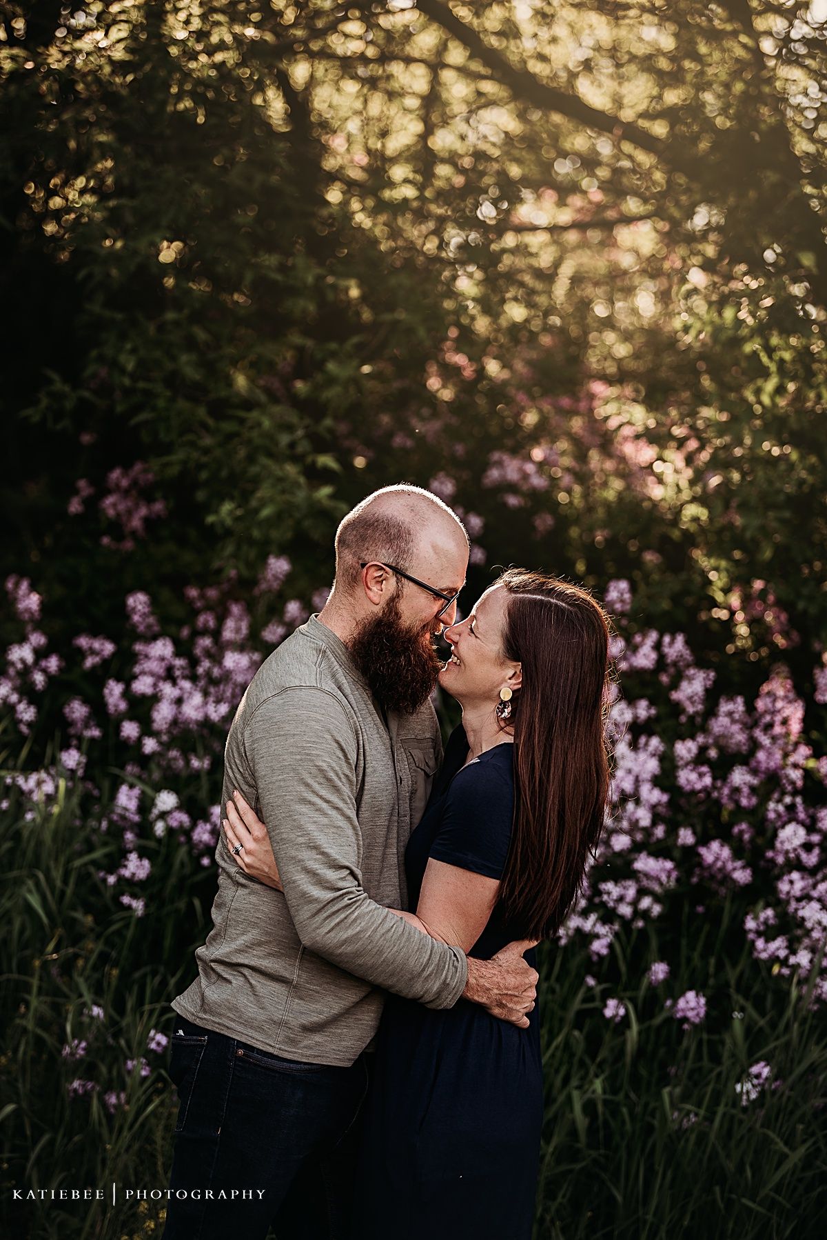 Romantic couple embrace in garden setting with purple flowers and golden sunset lighting.