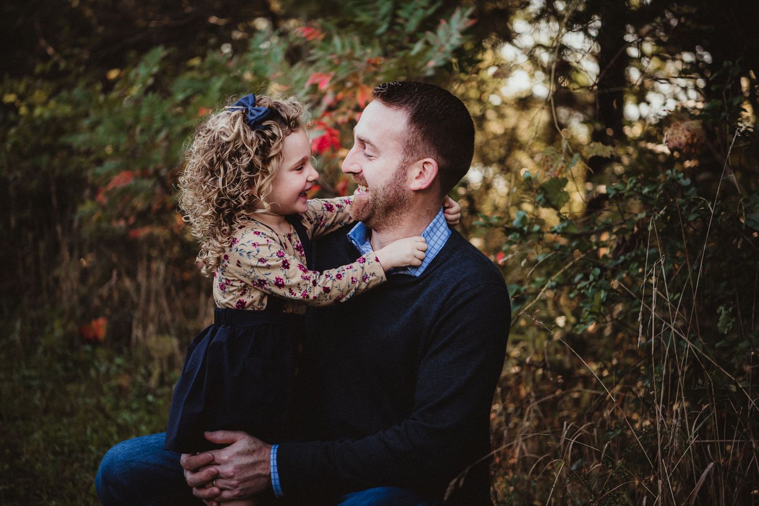 A heartwarming outdoor portrait shows a tender embrace between two people against a natural forest backdrop at sunset.