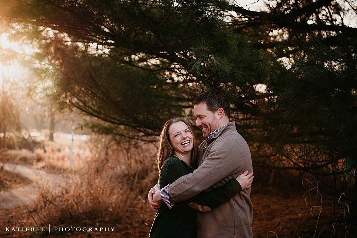 A romantic couple embraces outdoors during golden hour sunset with evergreen trees in the background.