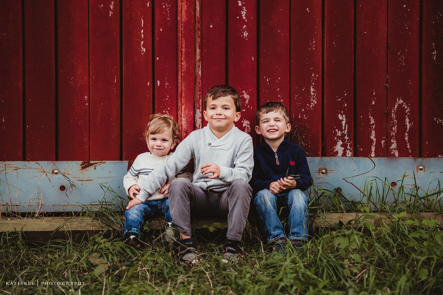 Three children sitting together on ground in front of weathered red barn.