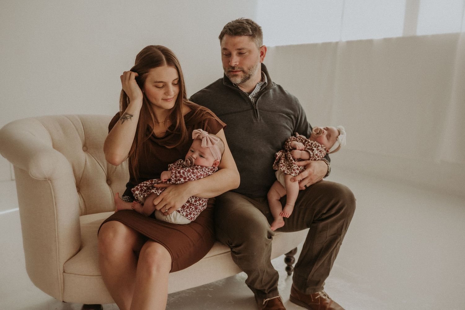 A family sits together on a beige couch holding twin babies in matching floral dresses.