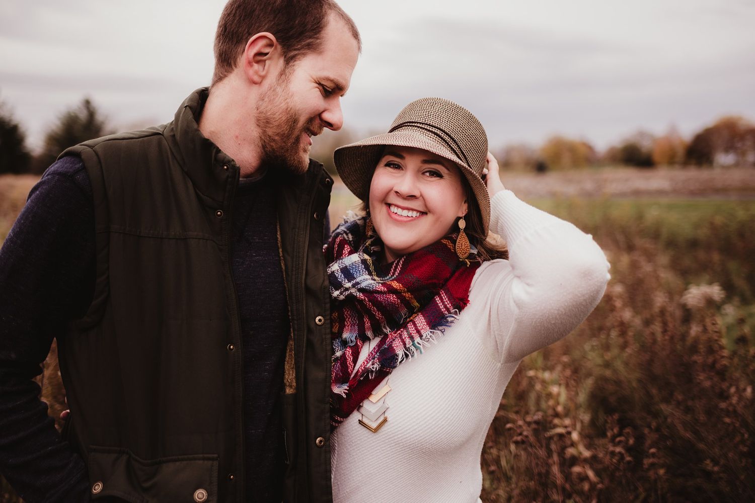 Couple sharing a joyful moment together in an autumn field, with her wearing a stylish hat and scarf.