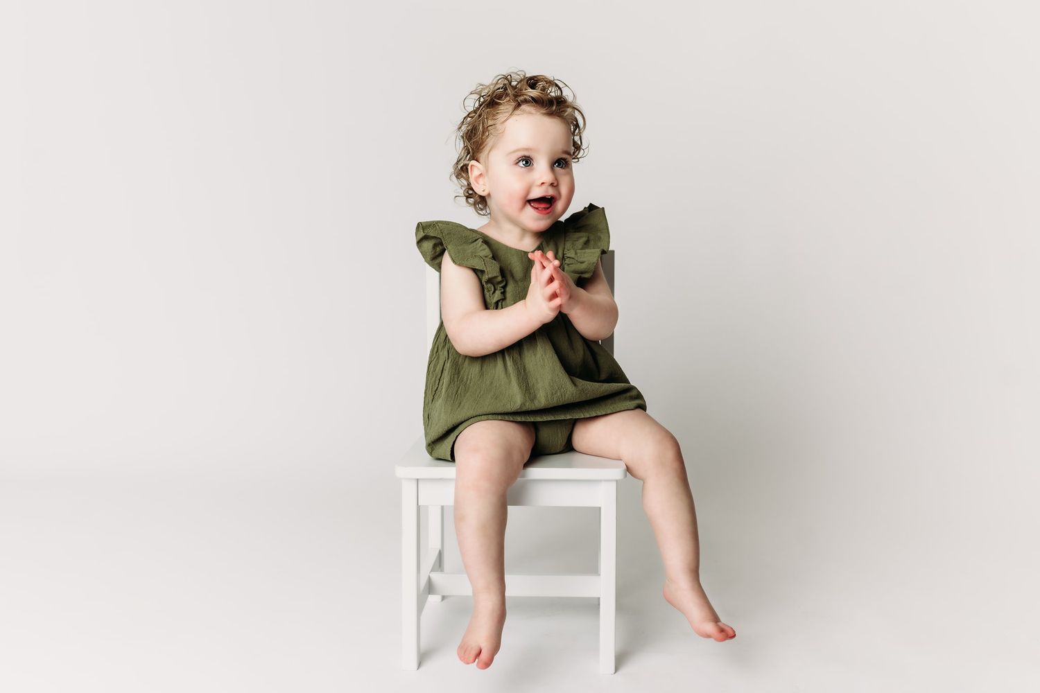 Child wearing olive dress sits happily on white chair in studio setting.