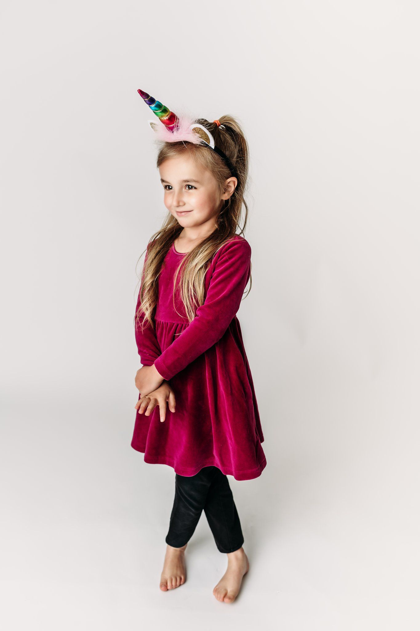 Child in red velvet dress and unicorn headband poses playfully on white background.