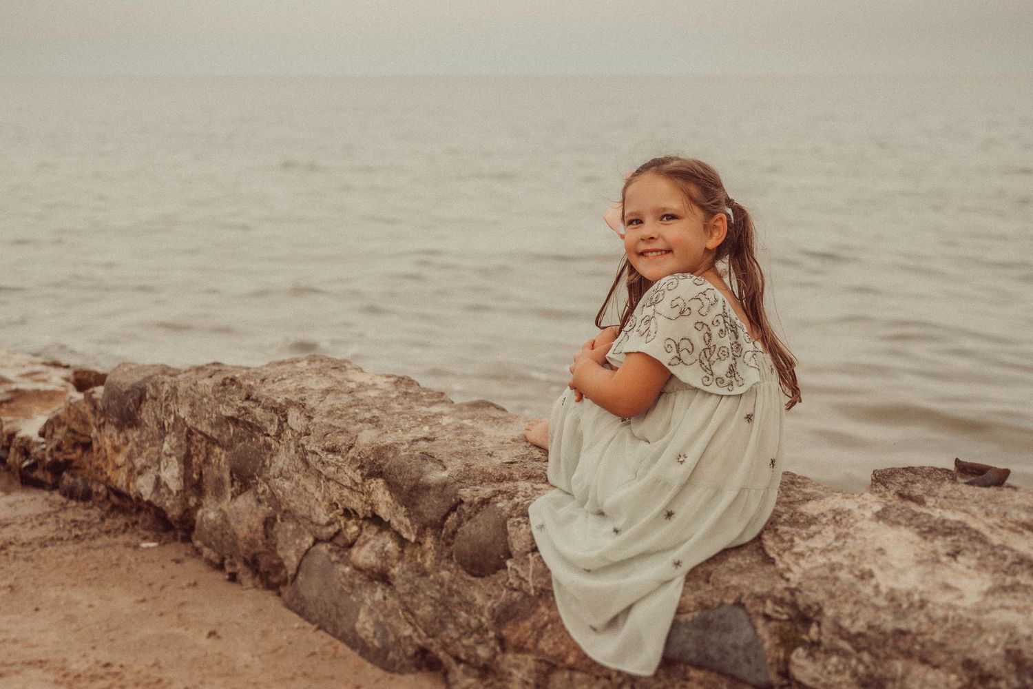 Child in vintage dress sits peacefully on rocky shore overlooking calm ocean waters.