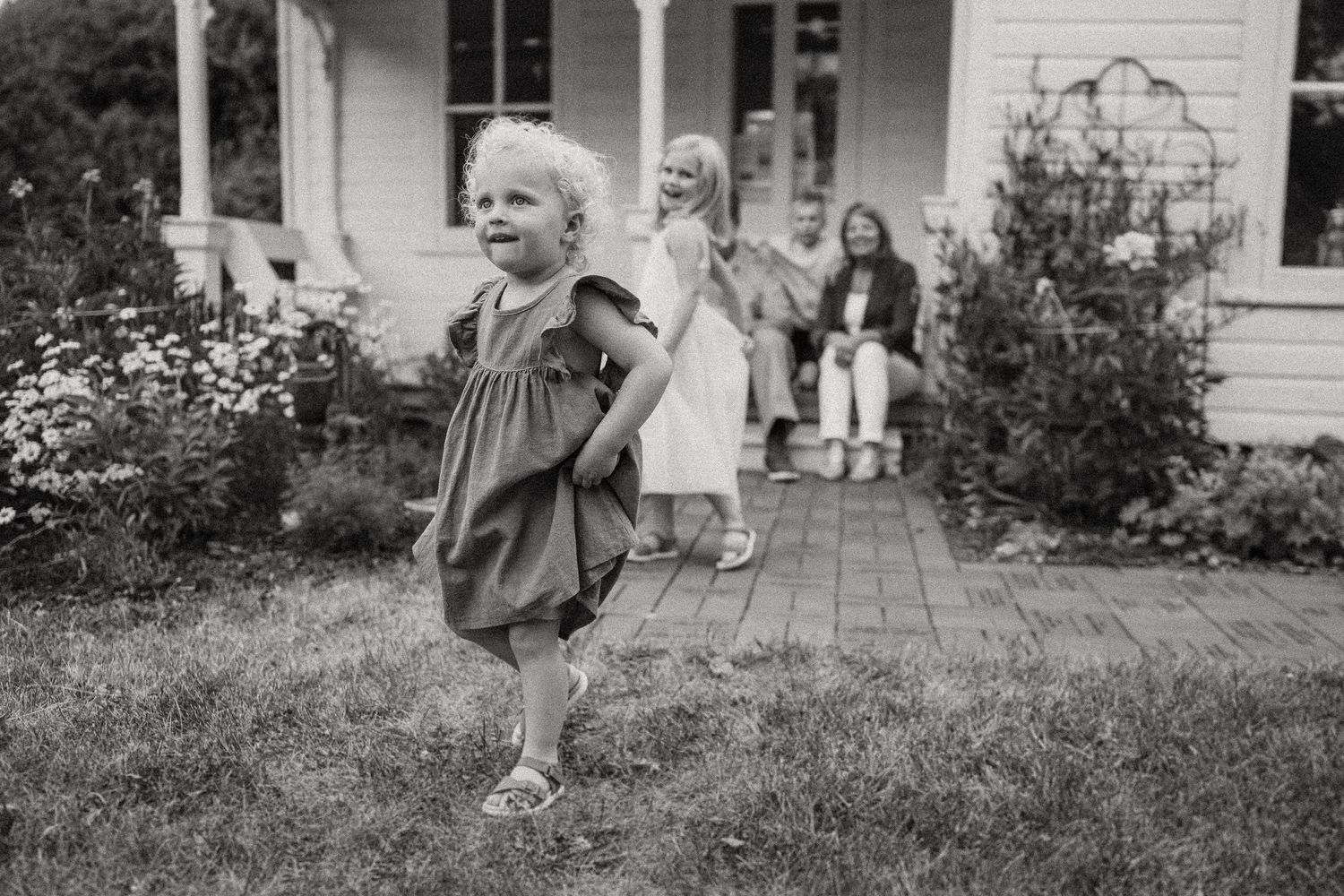 A toddler in a gray dress runs on a porch while others sit in the background.