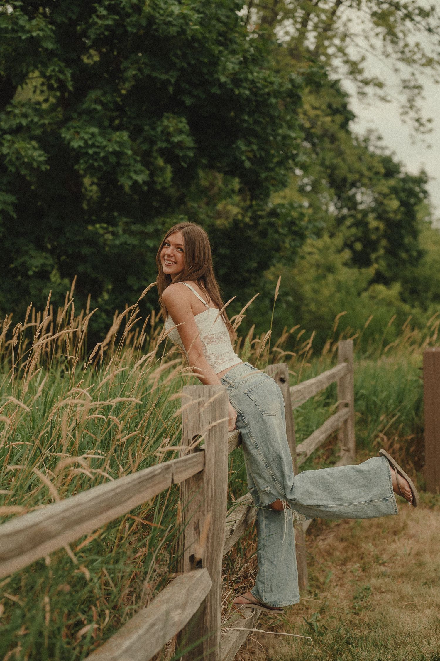Young woman in white top and wide-leg jeans leaning on rustic wooden fence surrounded by tall grass and trees.