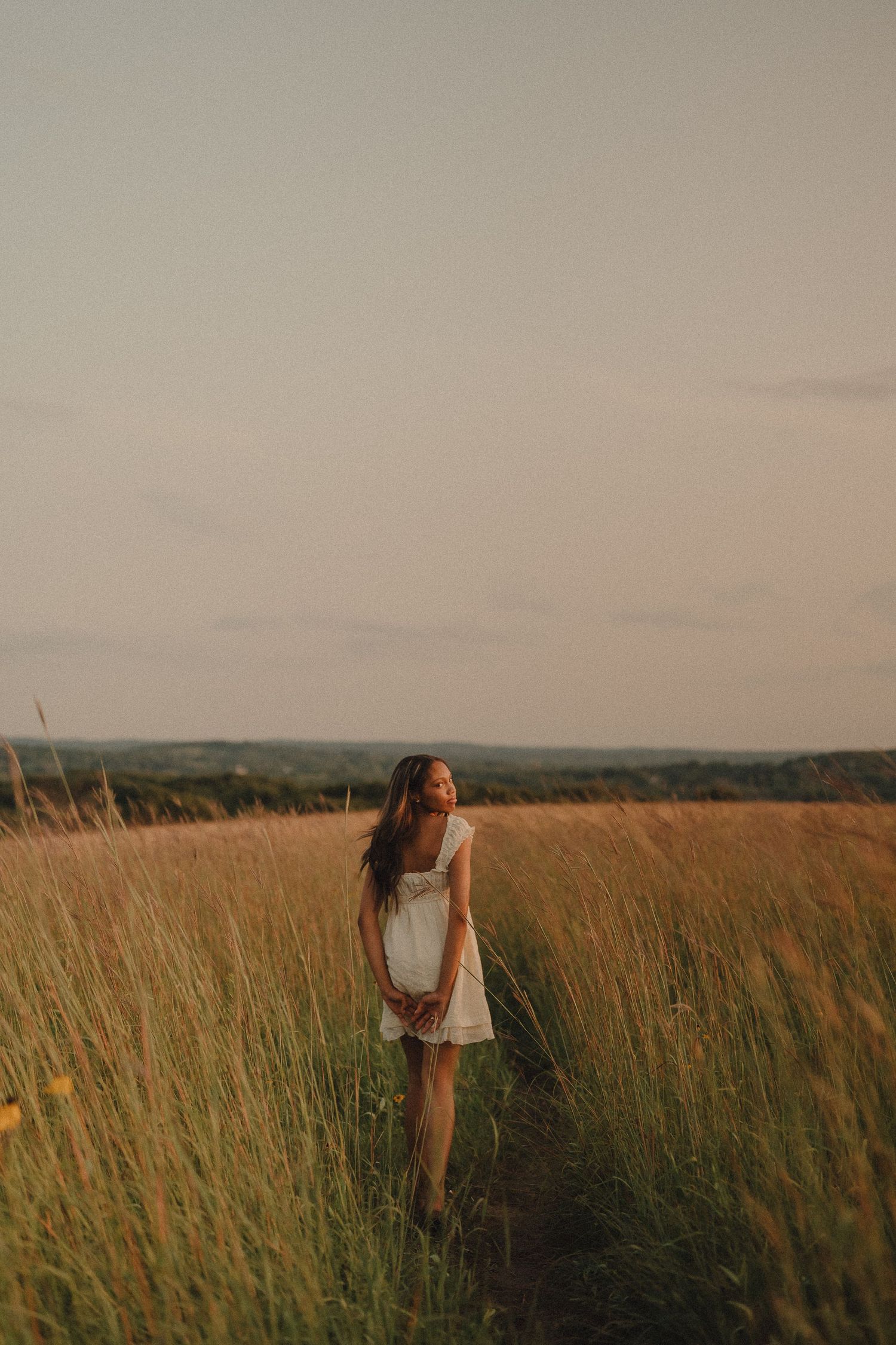 Young woman in white dress walking through tall golden grass field at sunset.