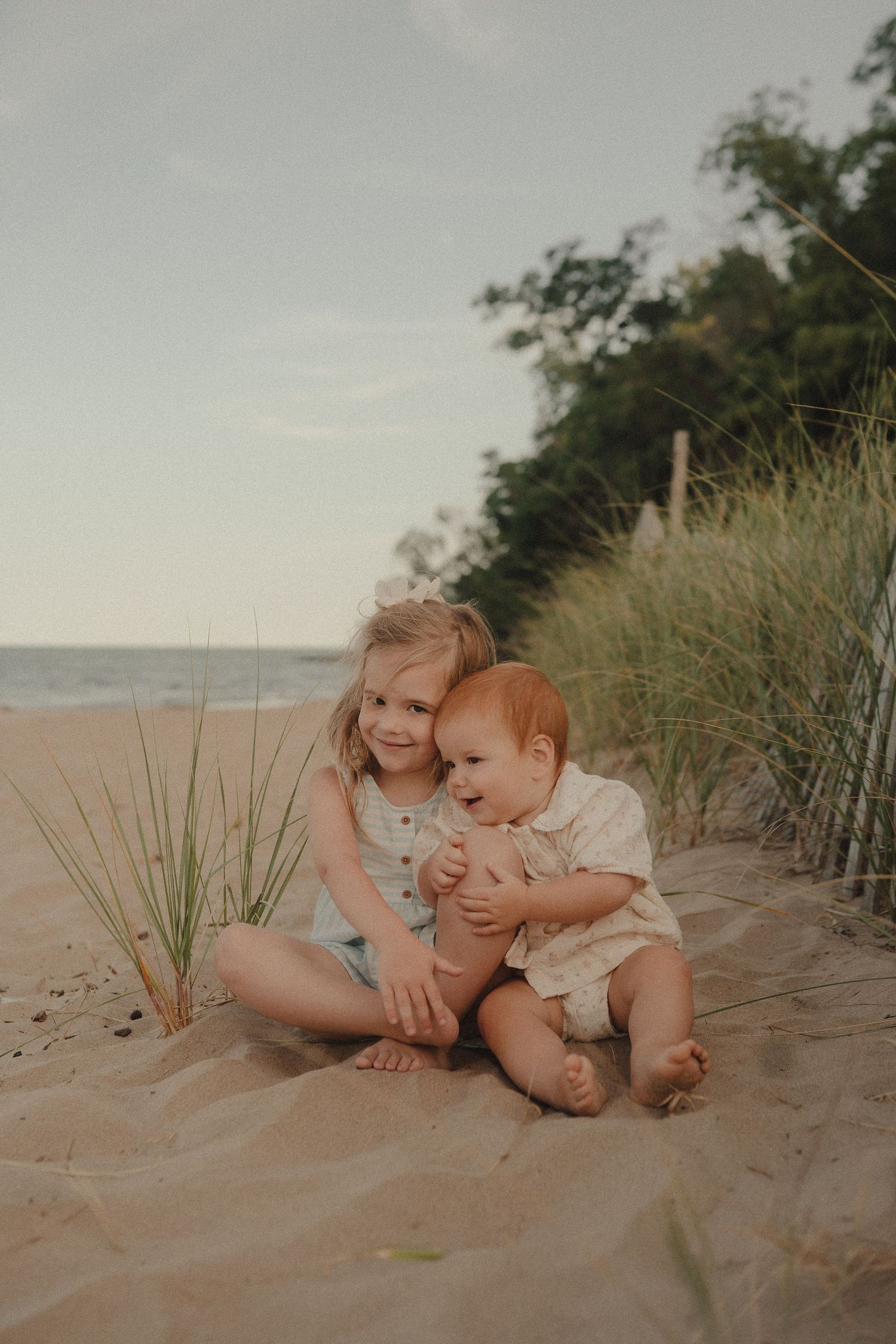 Two young siblings sit together on sandy beach, older girl hugging laughing red-haired toddler.