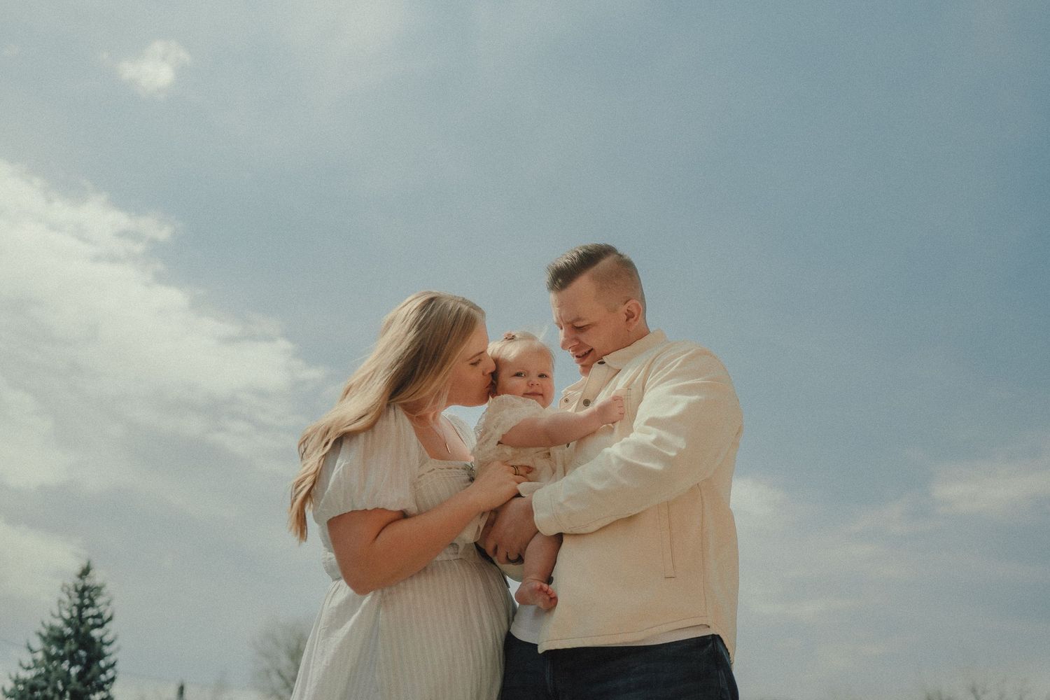 A happy family of three shares a tender moment outdoors under a blue sky with their infant baby.
