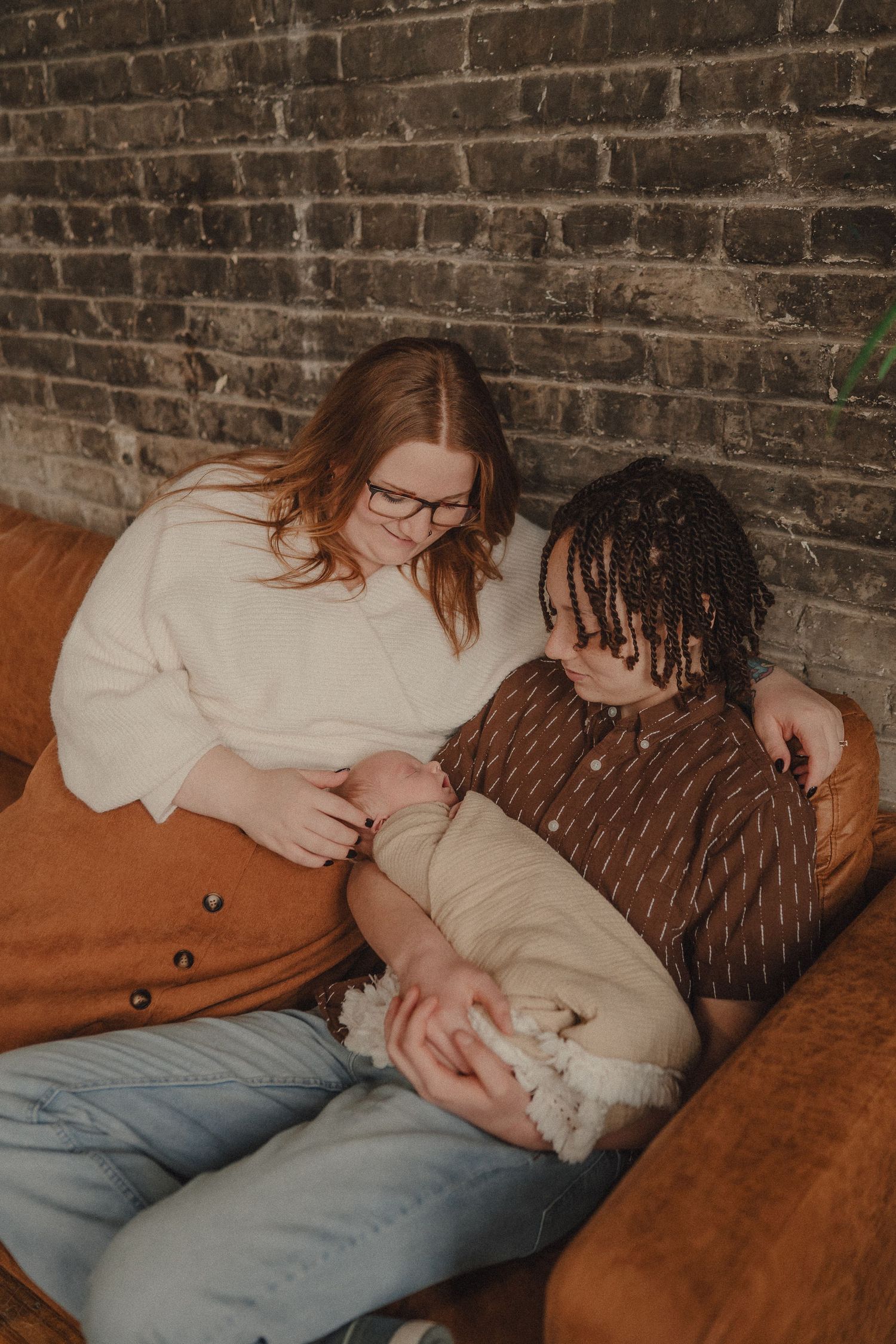 Two women sitting close together on a brown leather sofa against a brick wall, sharing a tender moment.