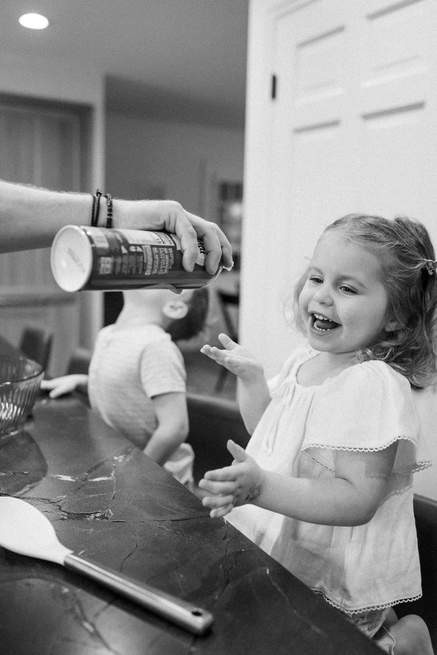 A laughing toddler girl in white dress enjoys baking with family at a flour-dusted kitchen counter.