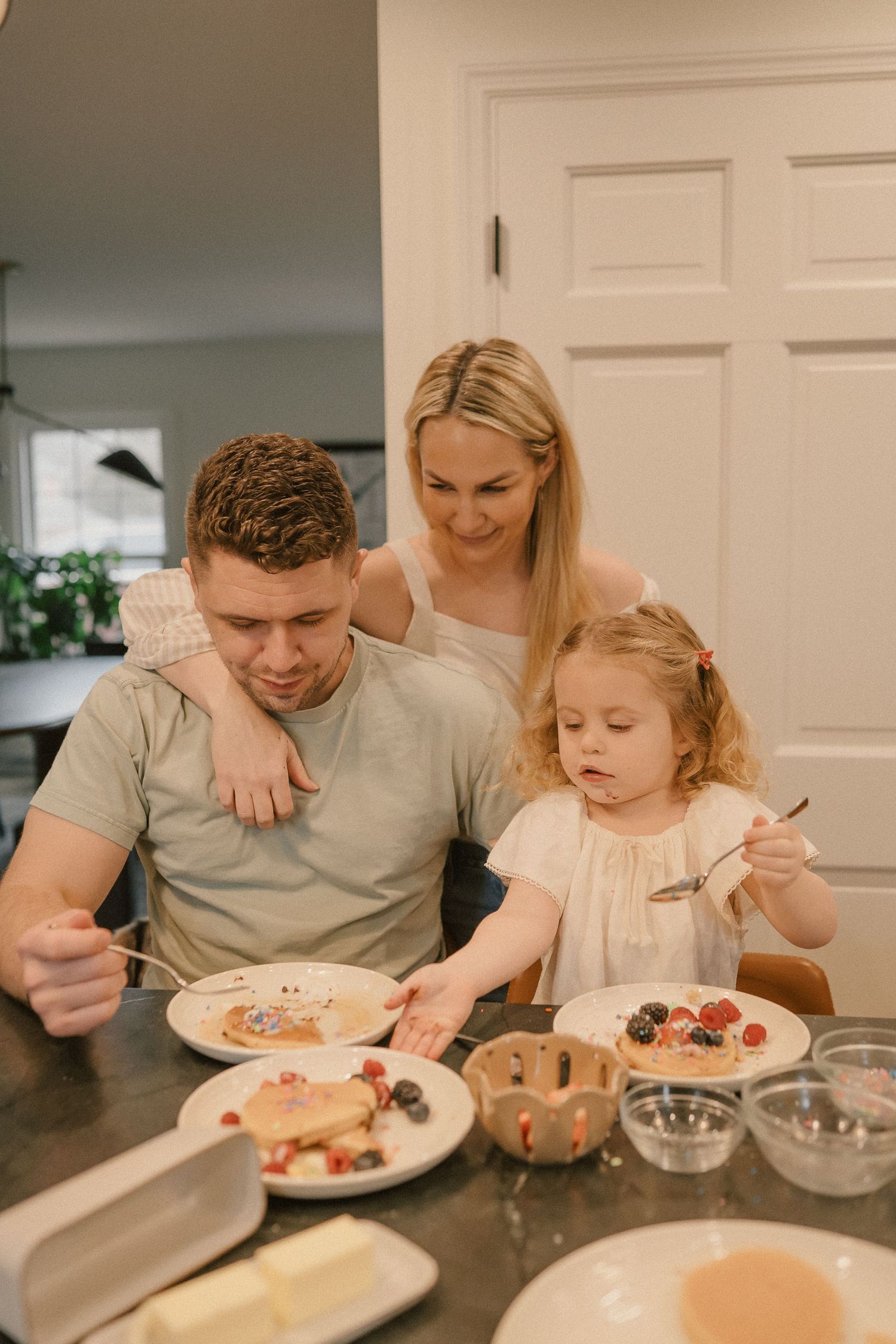 Family of three sharing pancake breakfast at a kitchen counter with berries and butter.